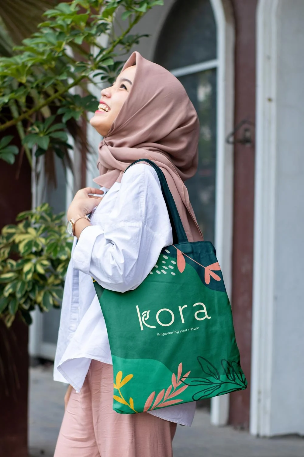 Woman holding a green Kora tote bag with pink, yellow, and dark green accents, featuring the beige Kora logo in the center.