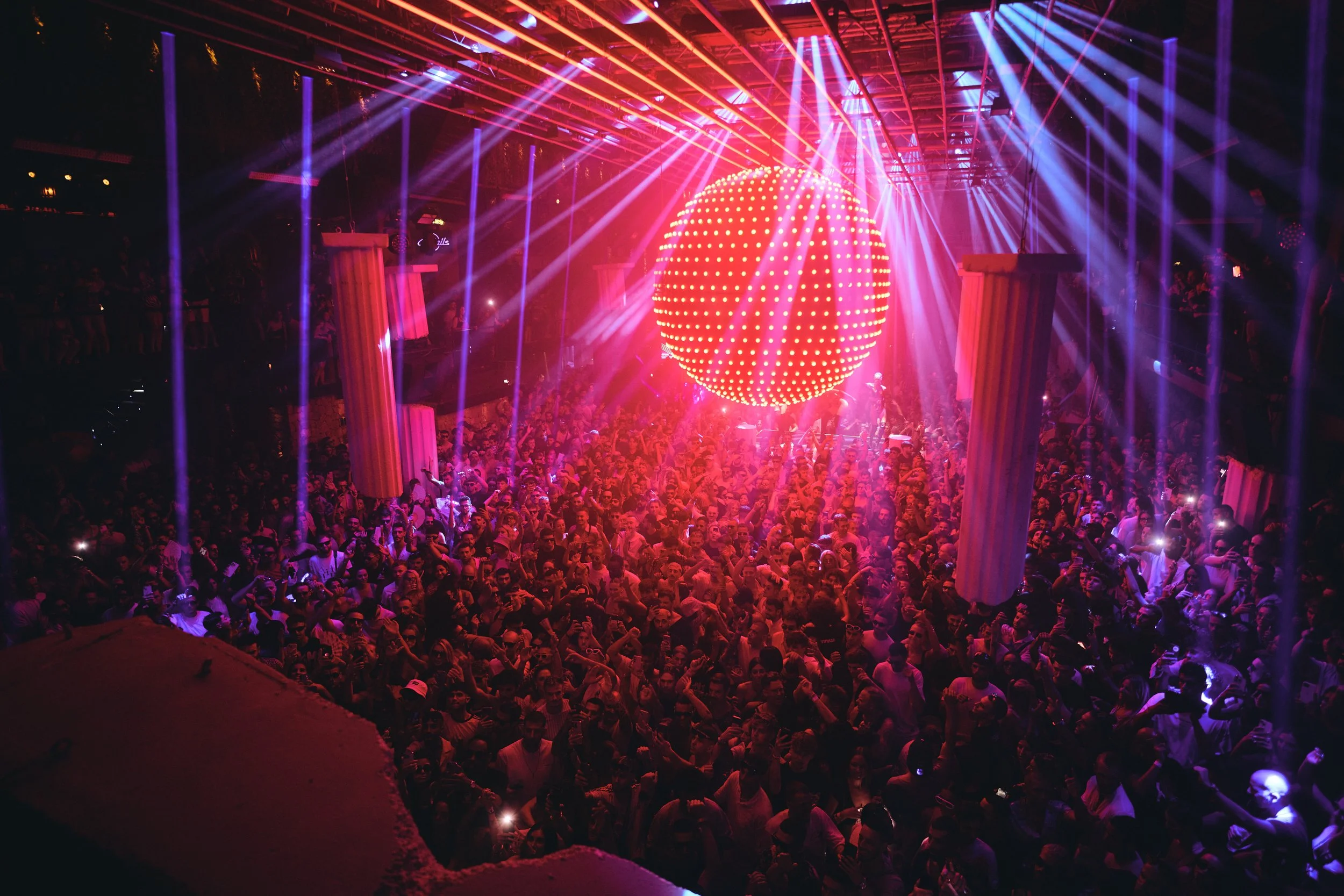 Packed nightclub in Ibiza with a crowd singing and dancing to Jamie Jones’ latest DJ set under a large pink disco ball, creating a vibrant party atmosphere.
