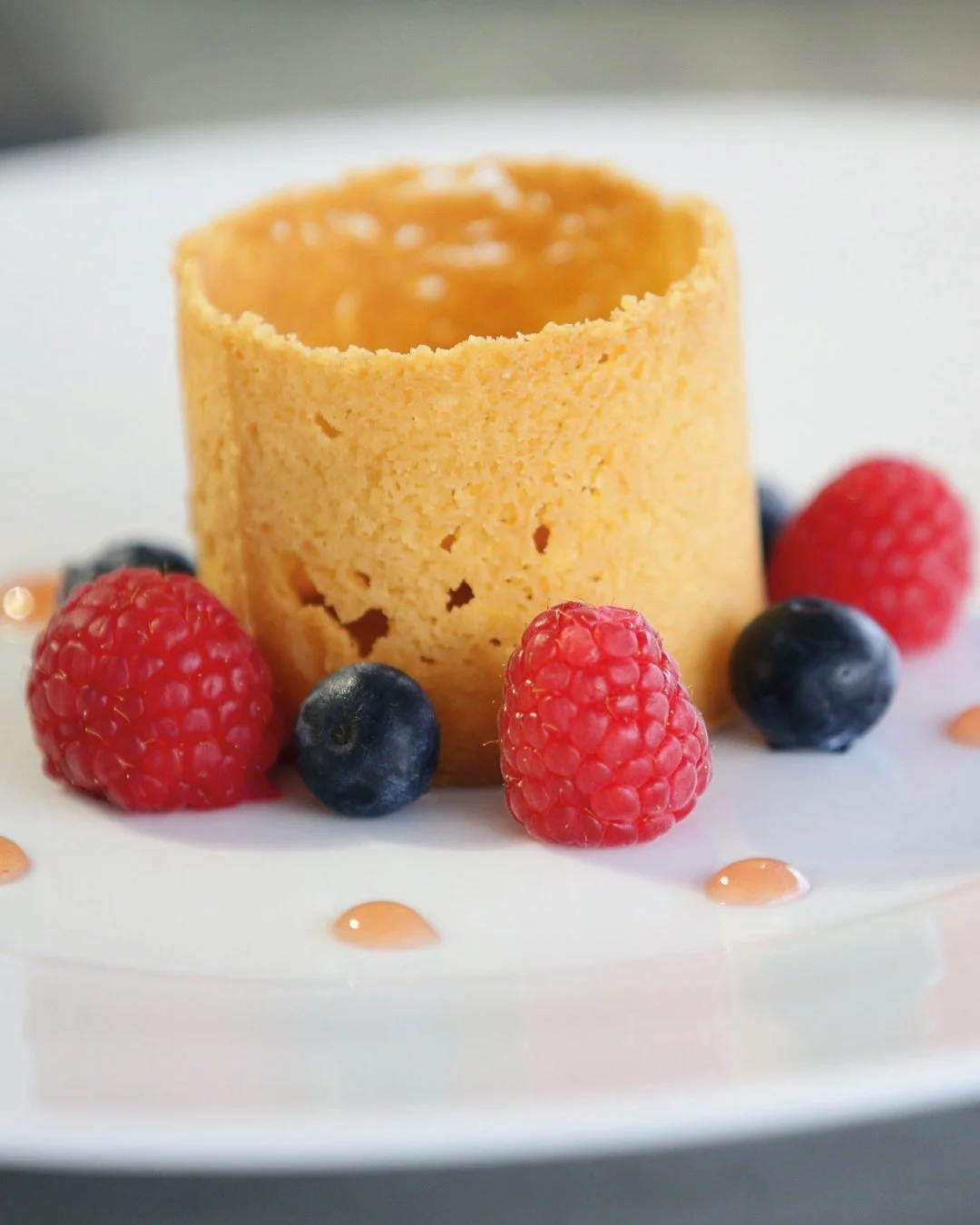 A dessert on a white plate featuring a round biscuit surrounded by vibrant raspberries and blueberries.