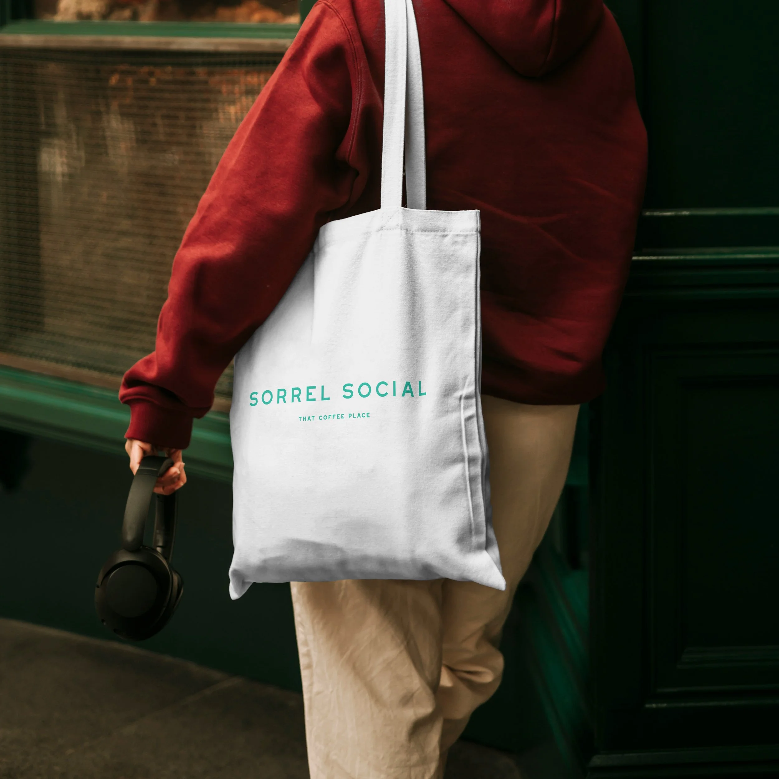 Person walking while carrying a white Sorrel Social tote bag over their shoulder. The bag features the Sorrel Social logo in green with the strapline ‘That Coffee Place’ underneath.
