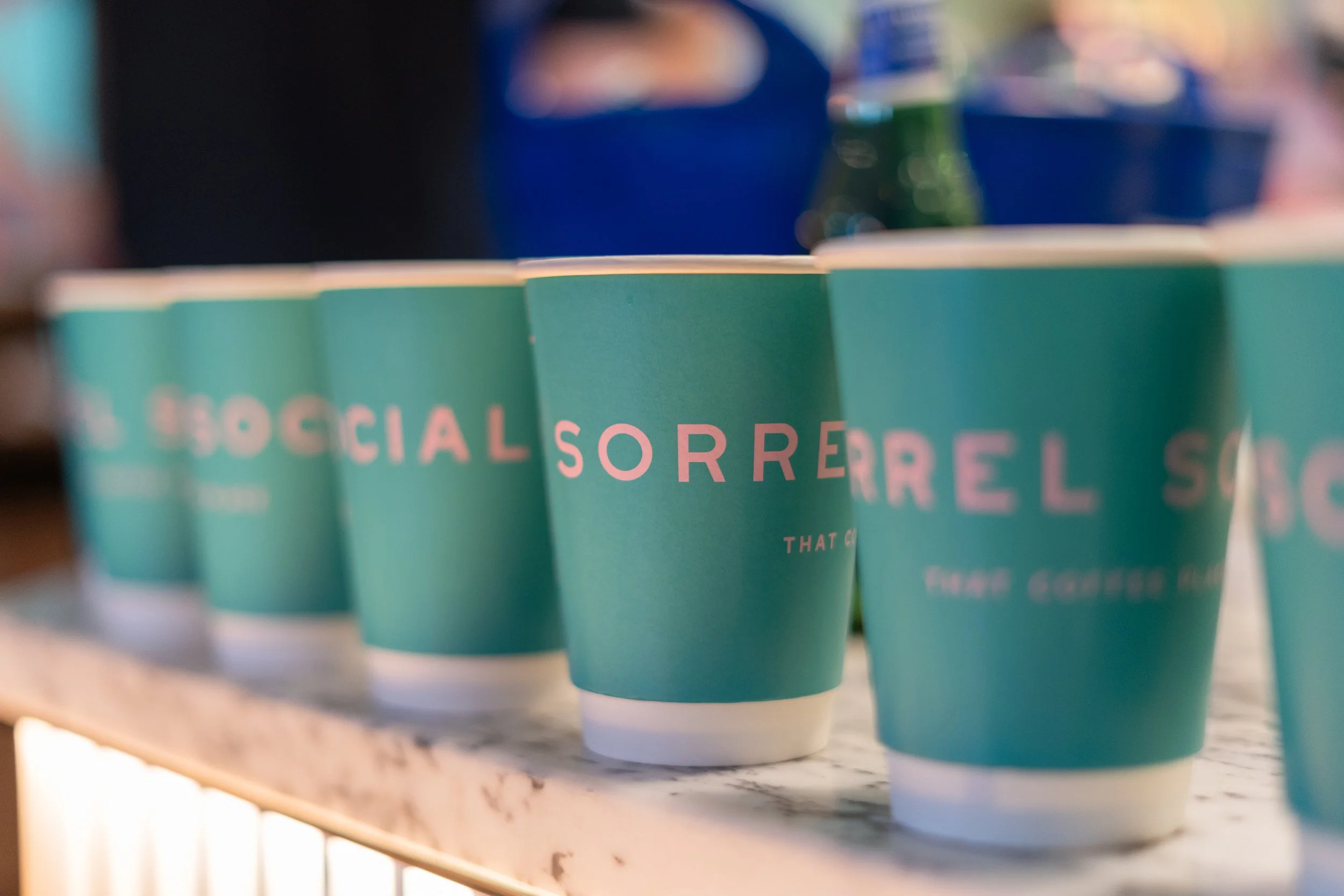 Sorrel Social green branded cups lined up on the counter, featuring a pink logo across the front.
