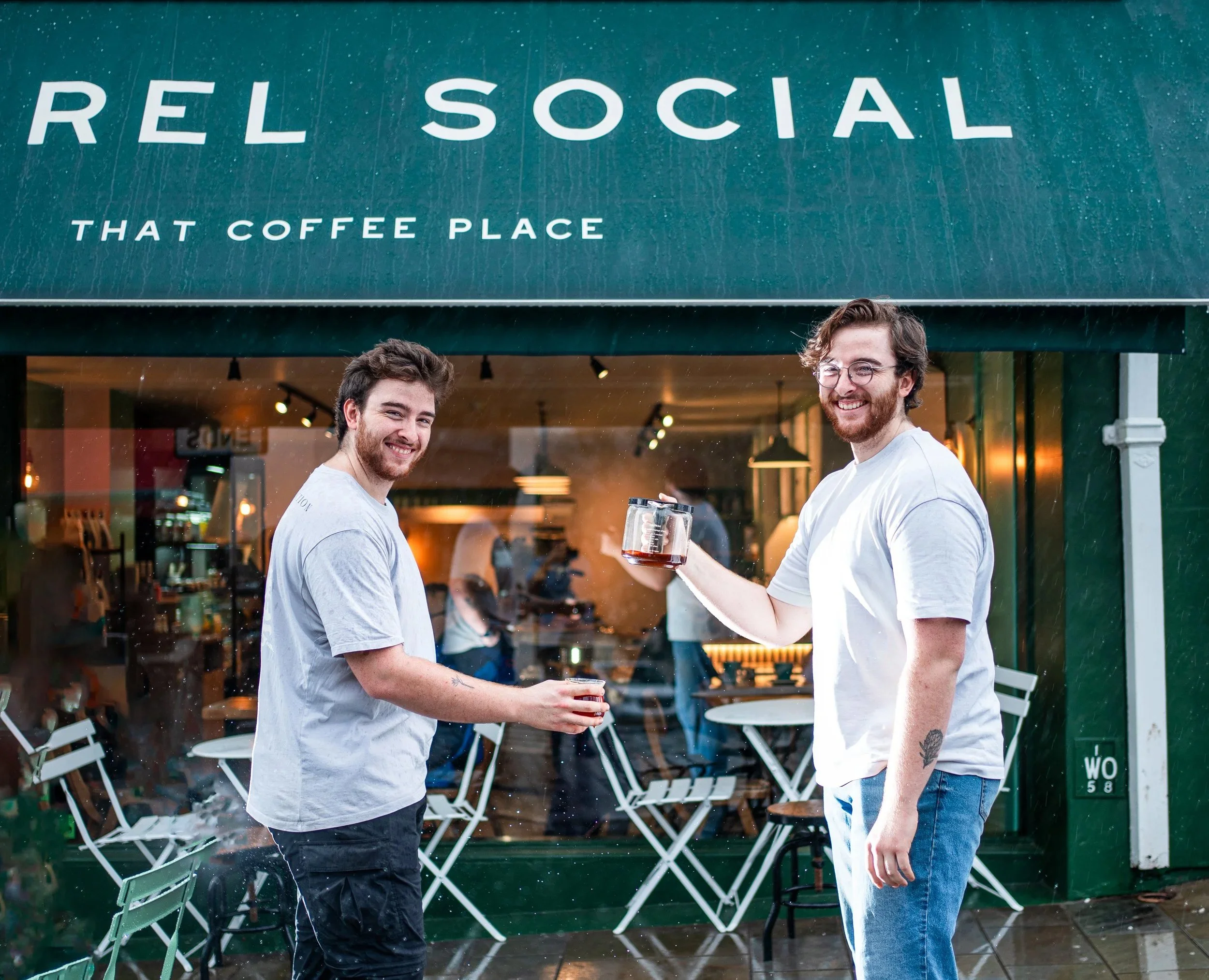 Two smiling baristas standing outside the Sorrel Social coffee shop, each holding a cup of coffee.