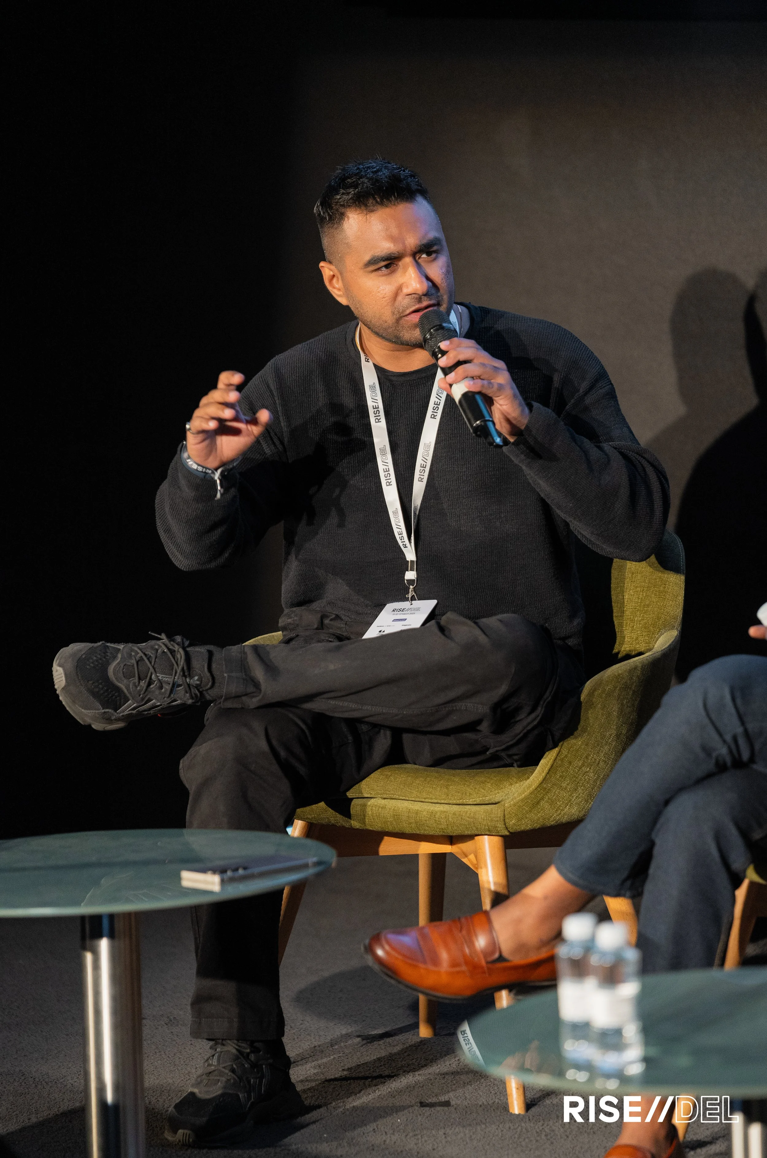Man speaking into microphone during panel discussion at RISE Festival, seated on a green chair with legs crossed, wearing a black sweater and black pants, with another speaker partly visible, and water bottles on tables in front.