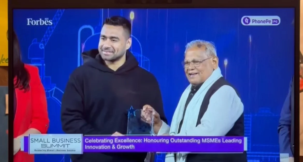 Two men shaking hands during an award presentation at a small business summit, with a blue background and event banners.