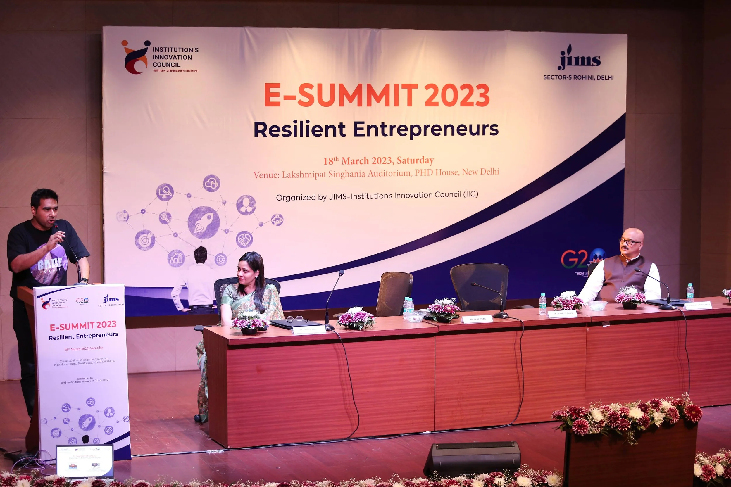 A man speaking at a podium on the left with a microphone, a woman sitting at a table with a floral arrangement in front of her, and a man sitting at the right side of the table, all in front of a large banner displaying the event details of E-SUMMIT 