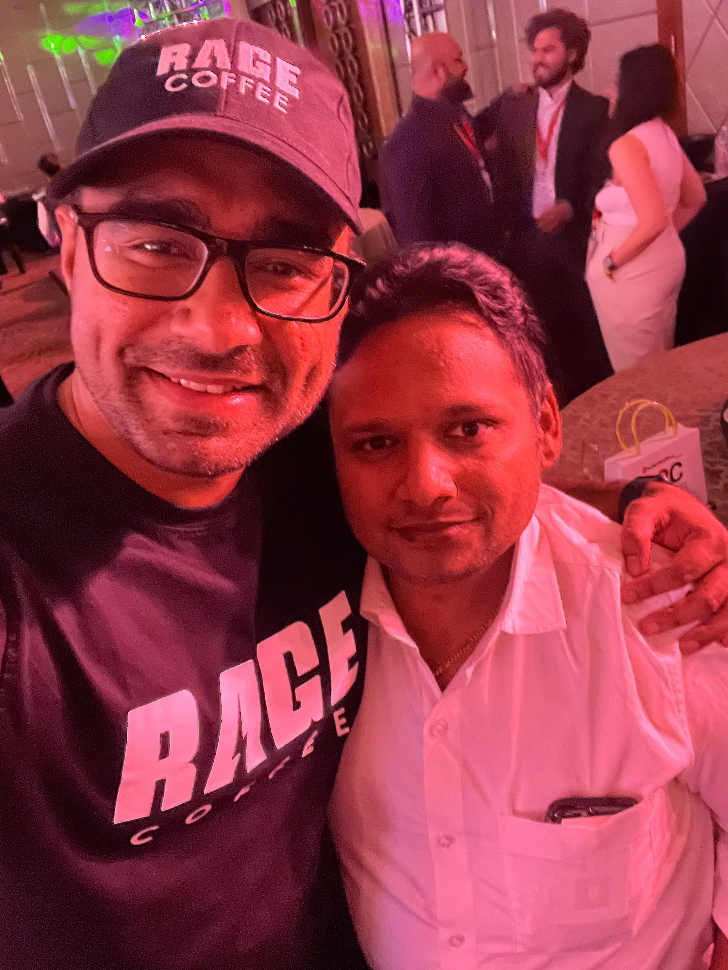 Two men taking a selfie at a social gathering or event, with one wearing a black cap and glasses, and the other in a white shirt. The background shows other people dressed formally, engaging in conversation, with dim lighting and colorful lights.