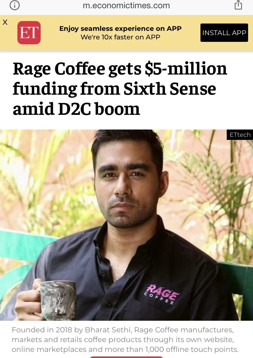 A man with dark hair and a beard holding a coffee mug, wearing a black shirt with 'RAGE' in pink and 'COFFEE' in white embroidered on the chest, sitting outdoors with greenery in the background.