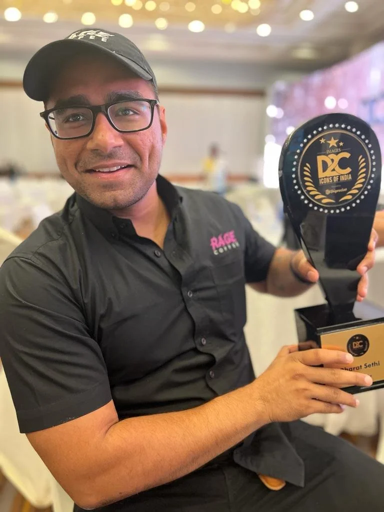 Man wearing black glasses, black cap, and black shirt holding a trophy with a circular logo. The background shows a banquet hall setting.