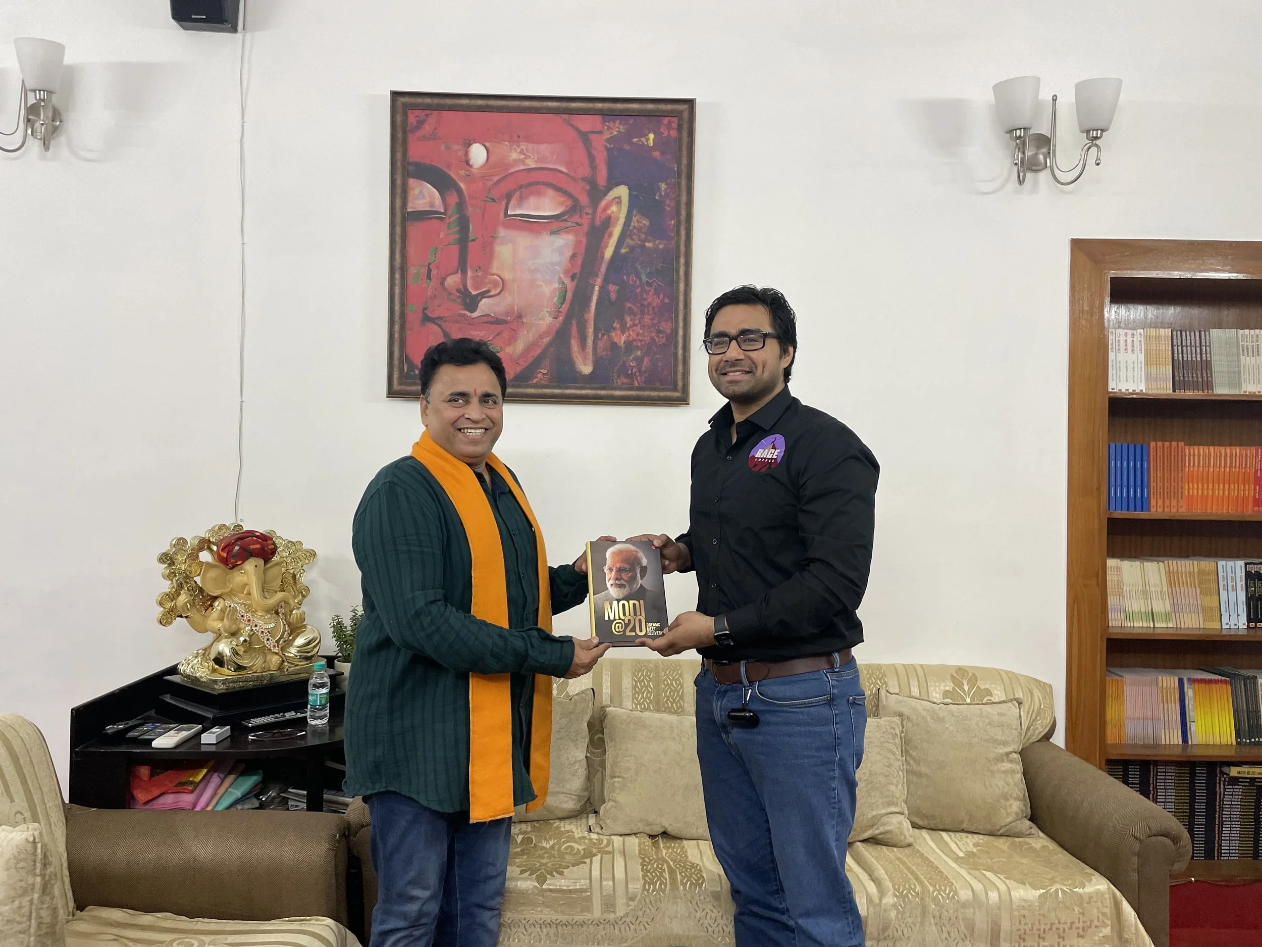 Two men standing in a living room, exchanging a book with an image of Narendra Modi on the cover. The man on the left wears a dark green kurta with an orange scarf, and the man on the right wears a black shirt and glasses. There is a sofa, a bookshel