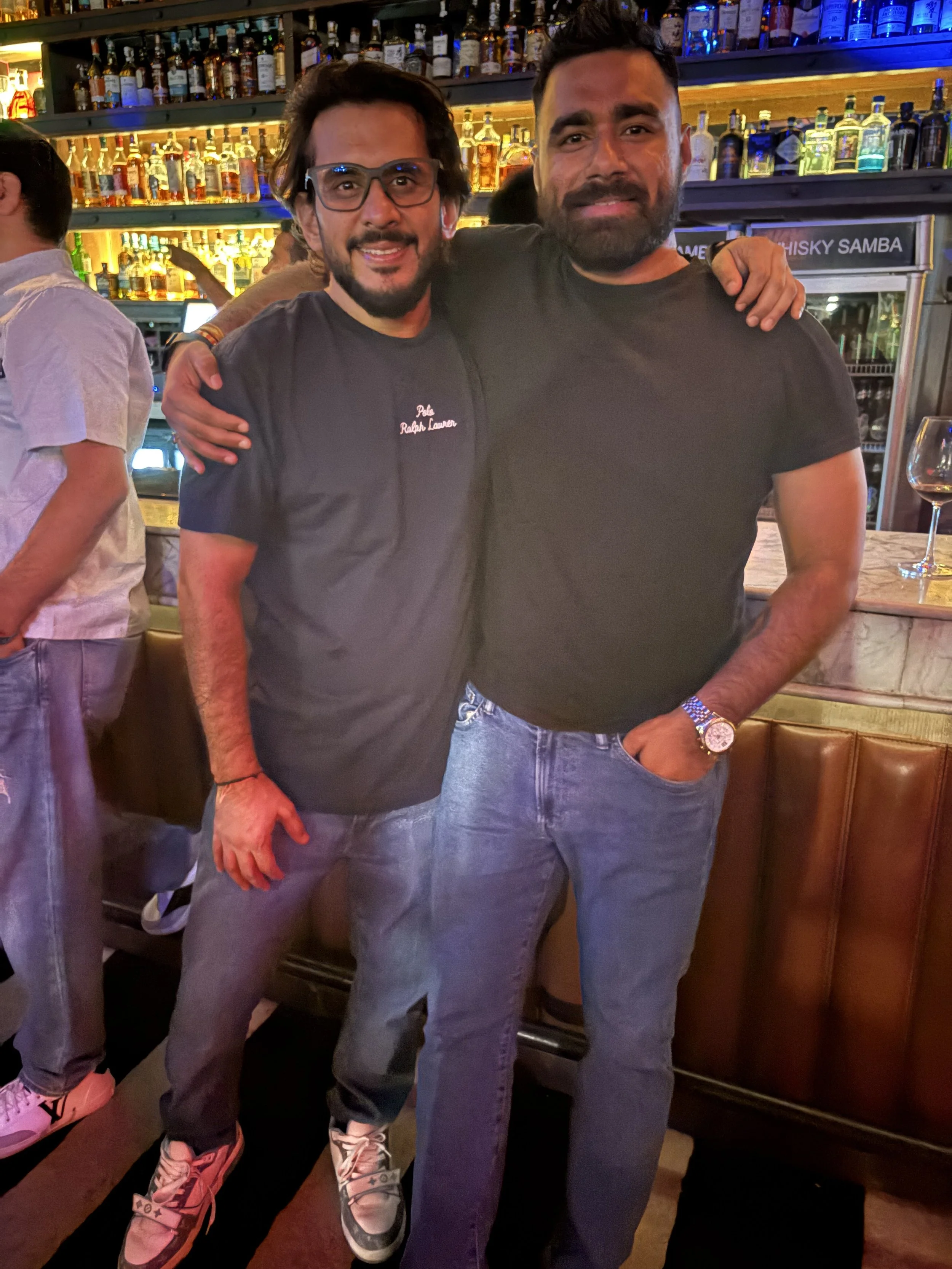 Two men standing side by side in a bar, smiling with arms around each other. The bar is stocked with various bottles of alcohol and there is a wine glass on the counter to the right.