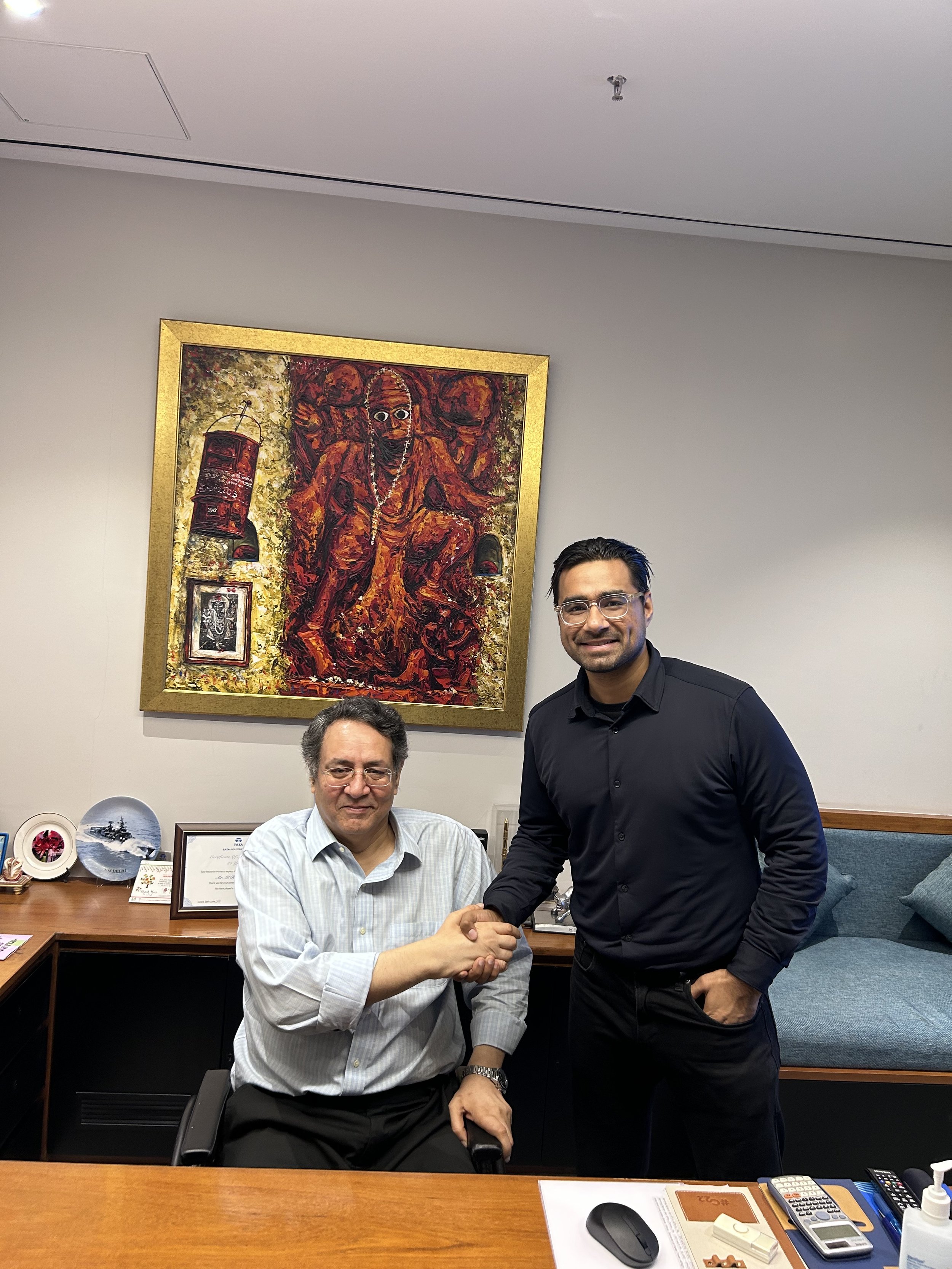 Two men shaking hands in an office with a large colorful painting on the wall behind them. One man is sitting at a desk and the other is standing.