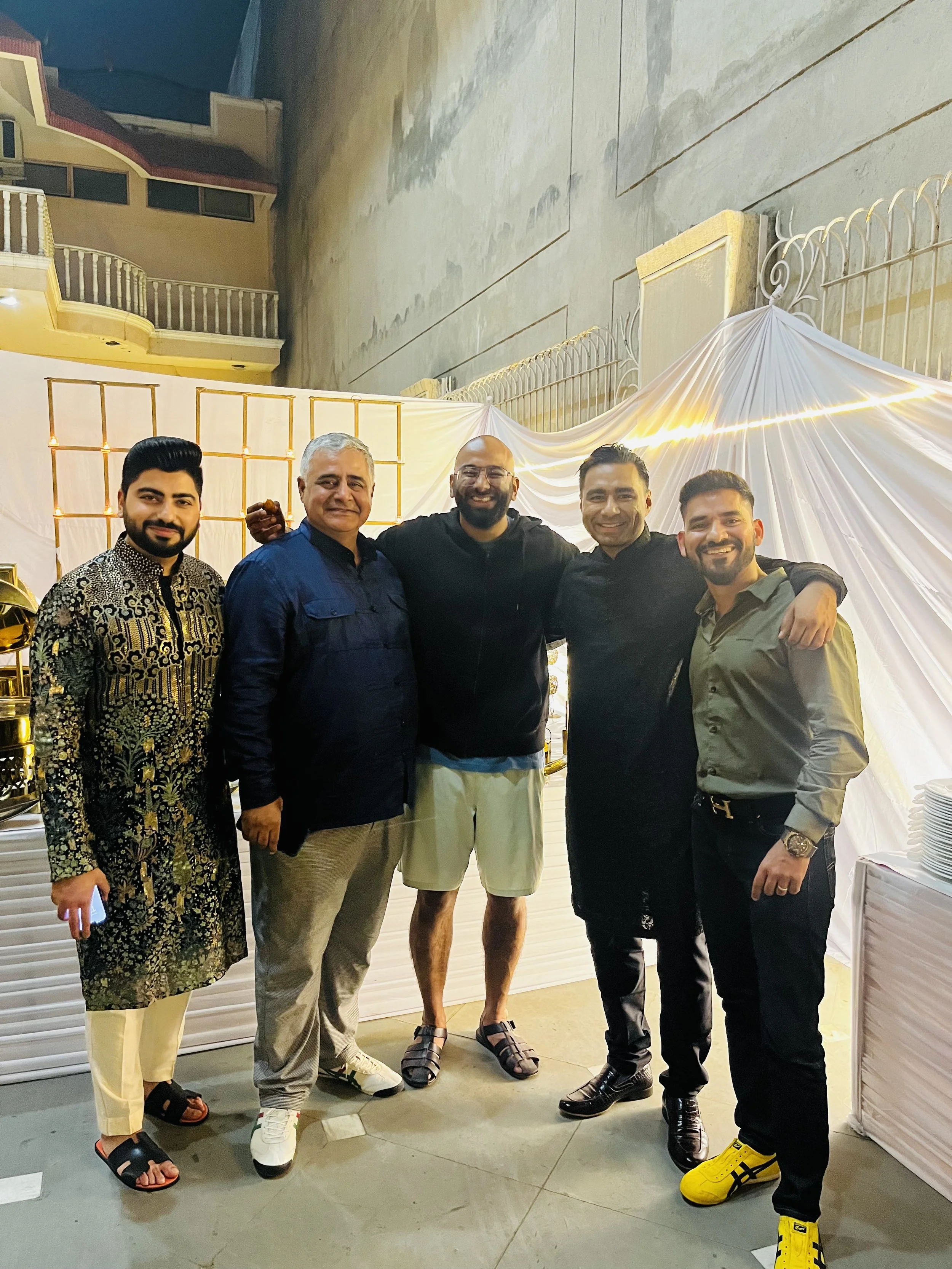 Group of five men standing together at an outdoor celebration or event, with a decorated backdrop of white drapery and lights behind them.