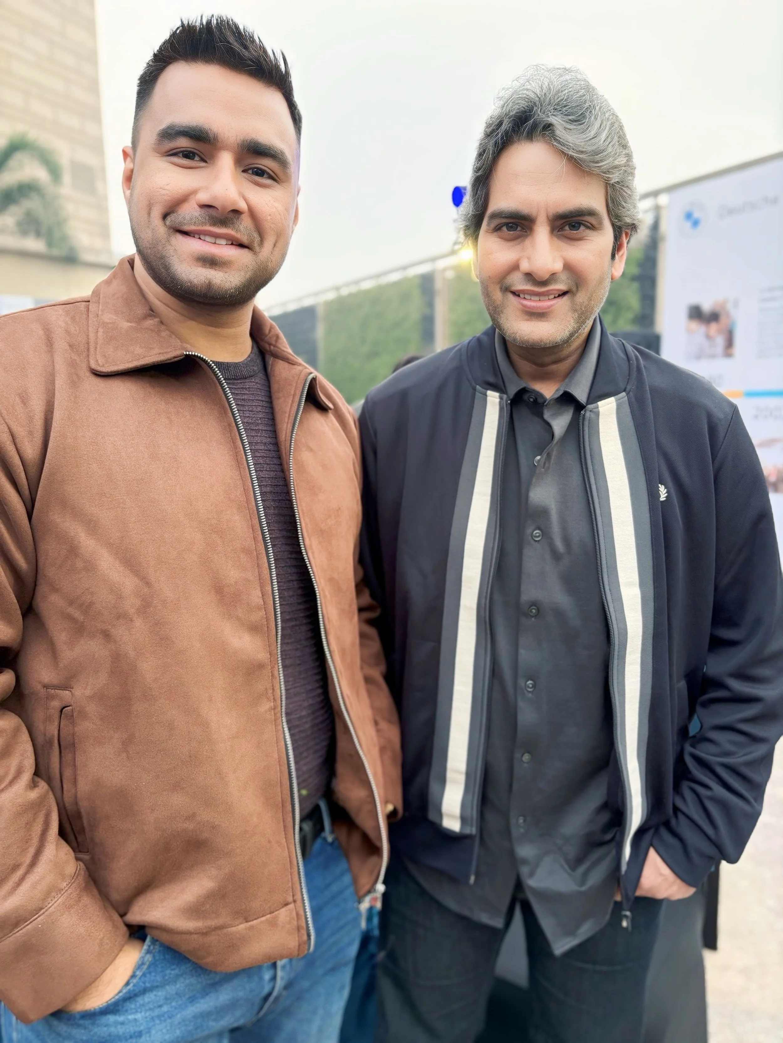 Two men smiling outdoors with a banner and some greenery in the background.