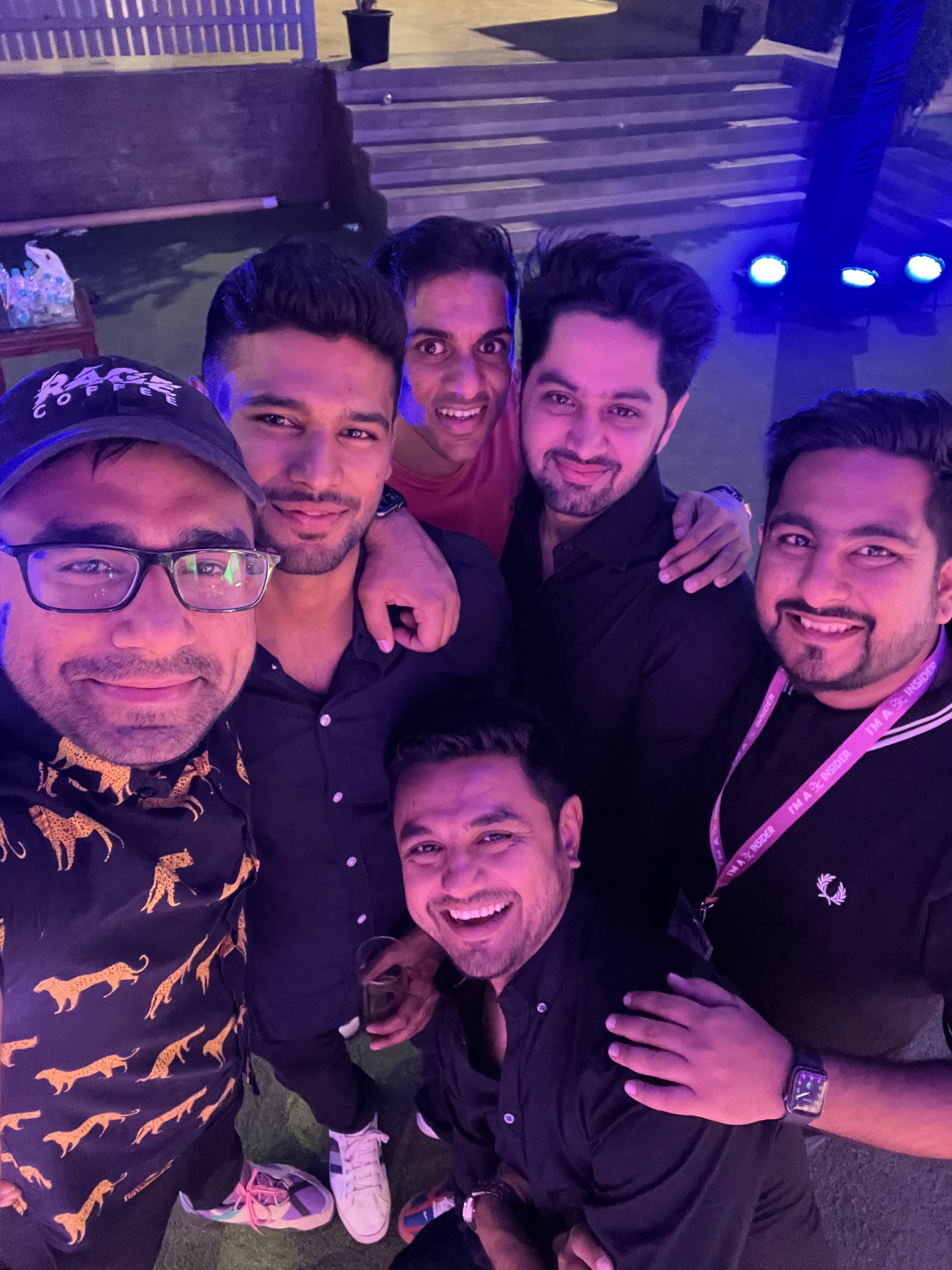 Group of six men smiling and taking a selfie outdoors at night with purple lighting, steps in the background, and decorative plants.