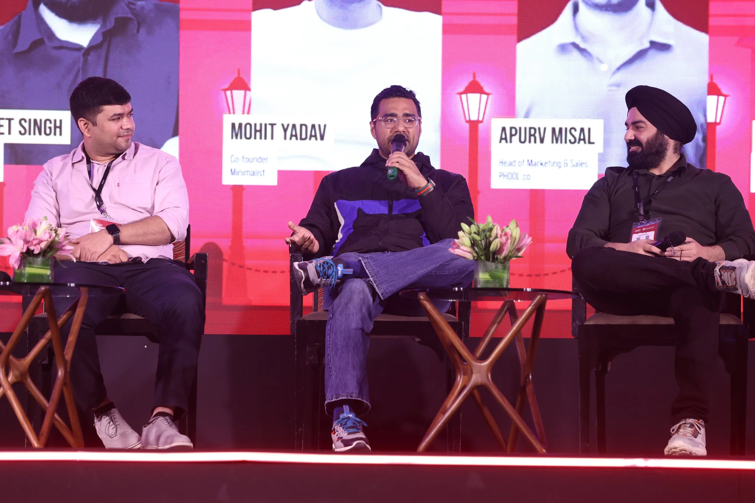Three men seated on a stage during a panel discussion. The man in the center is speaking into a microphone, wearing glasses, a black jacket with blue accents, and jeans. The man on the left wears a light pink shirt, dark pants, and white sneakers. Th