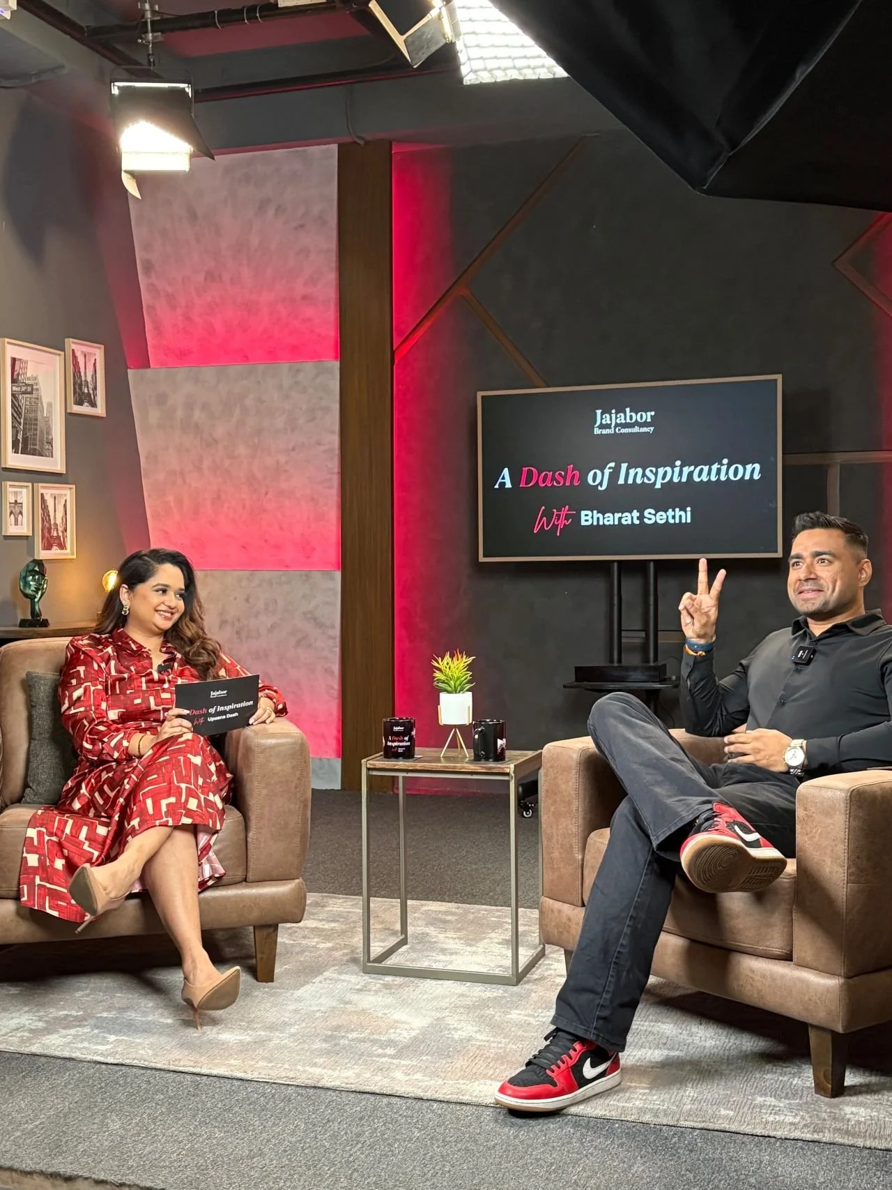 TV talk show set with a woman in a red patterned outfit sitting on a sofa, a man in casual black and red sneakers sitting on a chair, and a screen behind them displaying the show title 'A Dash of Inspiration with Bharat Sethi' and the guest 'Jabbar B