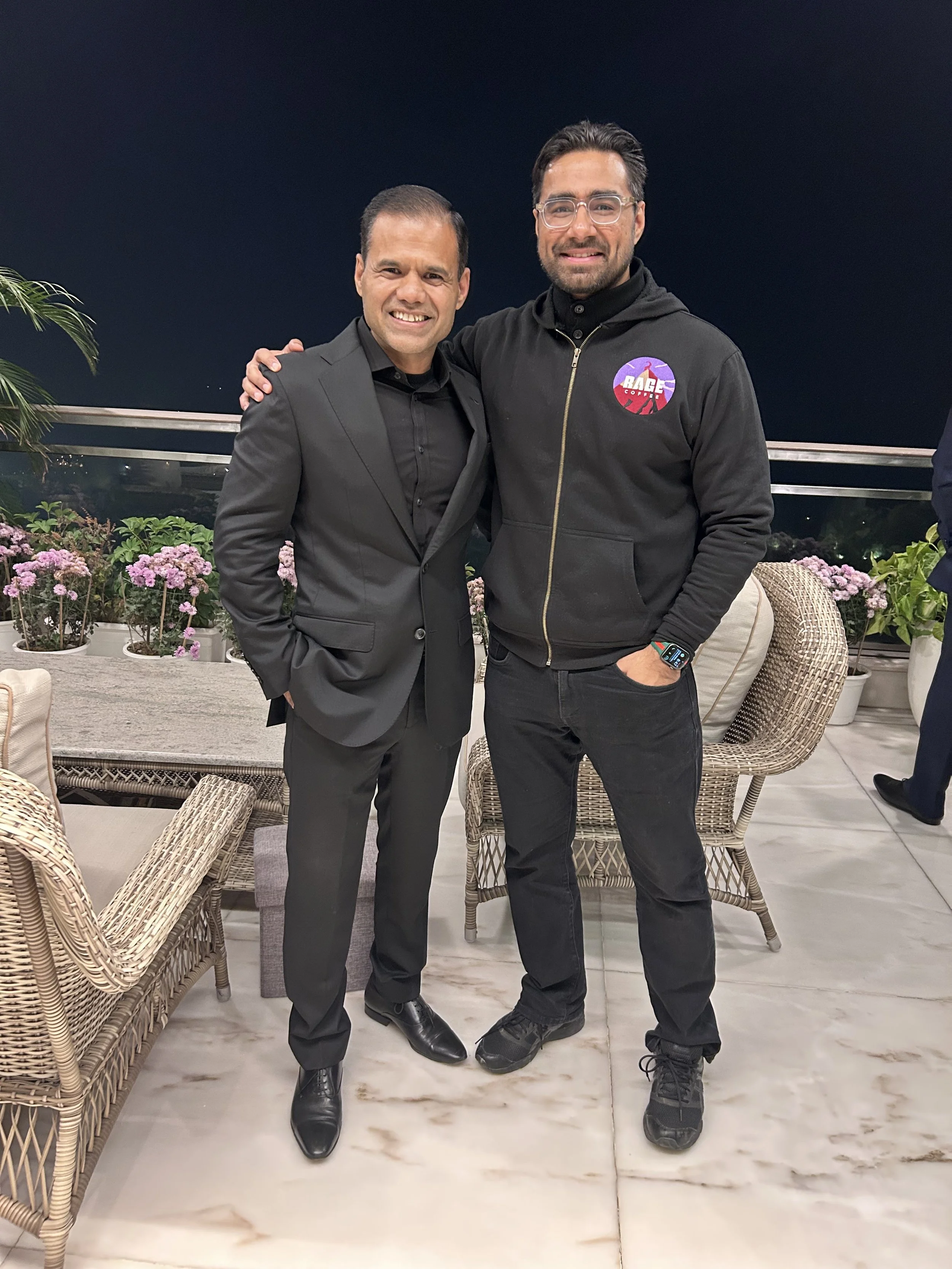 Two men standing together at night, smiling, with a balcony and flowers in the background.