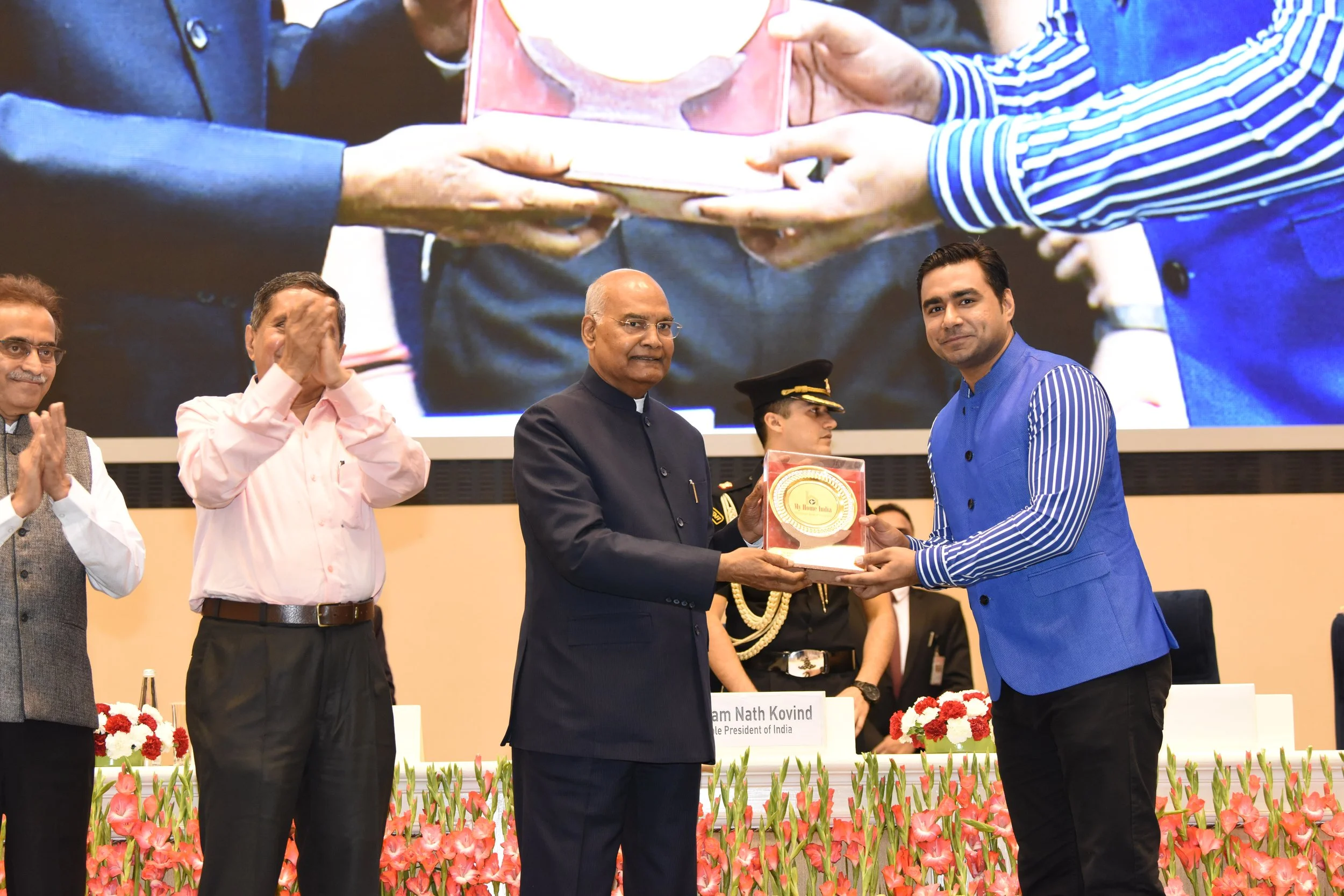 Indian President Droupadi Murmu presenting an award to a young man in a blue vest during a formal ceremony, with other officials and uniformed personnel on stage and a large screen showing a close-up of the award presentation in the background, decor