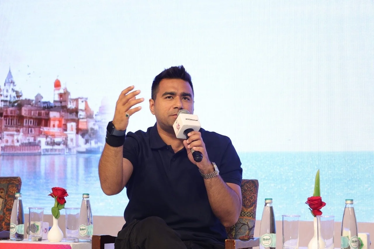 A man speaking into a microphone during a panel discussion, seated at a table with water bottles and vases with red roses, with a backdrop of a digital ocean scene and buildings.