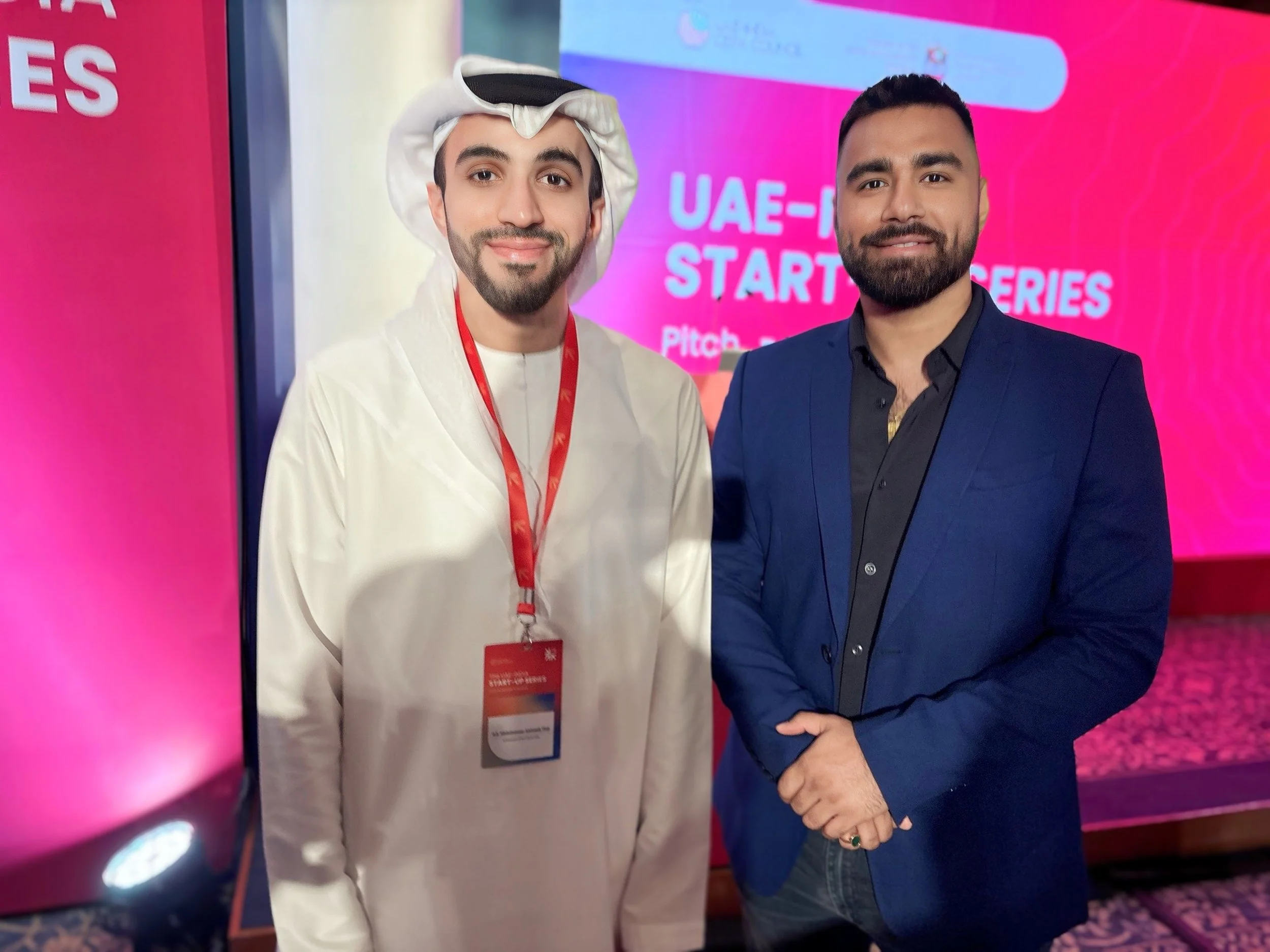 Two men standing in front of a pink and purple backdrop that reads 'UAE-H STARTUP SERIES'. One man is dressed in traditional Middle Eastern attire, wearing a white thobe and head covering, with a conference badge, and the other is wearing a dark blue