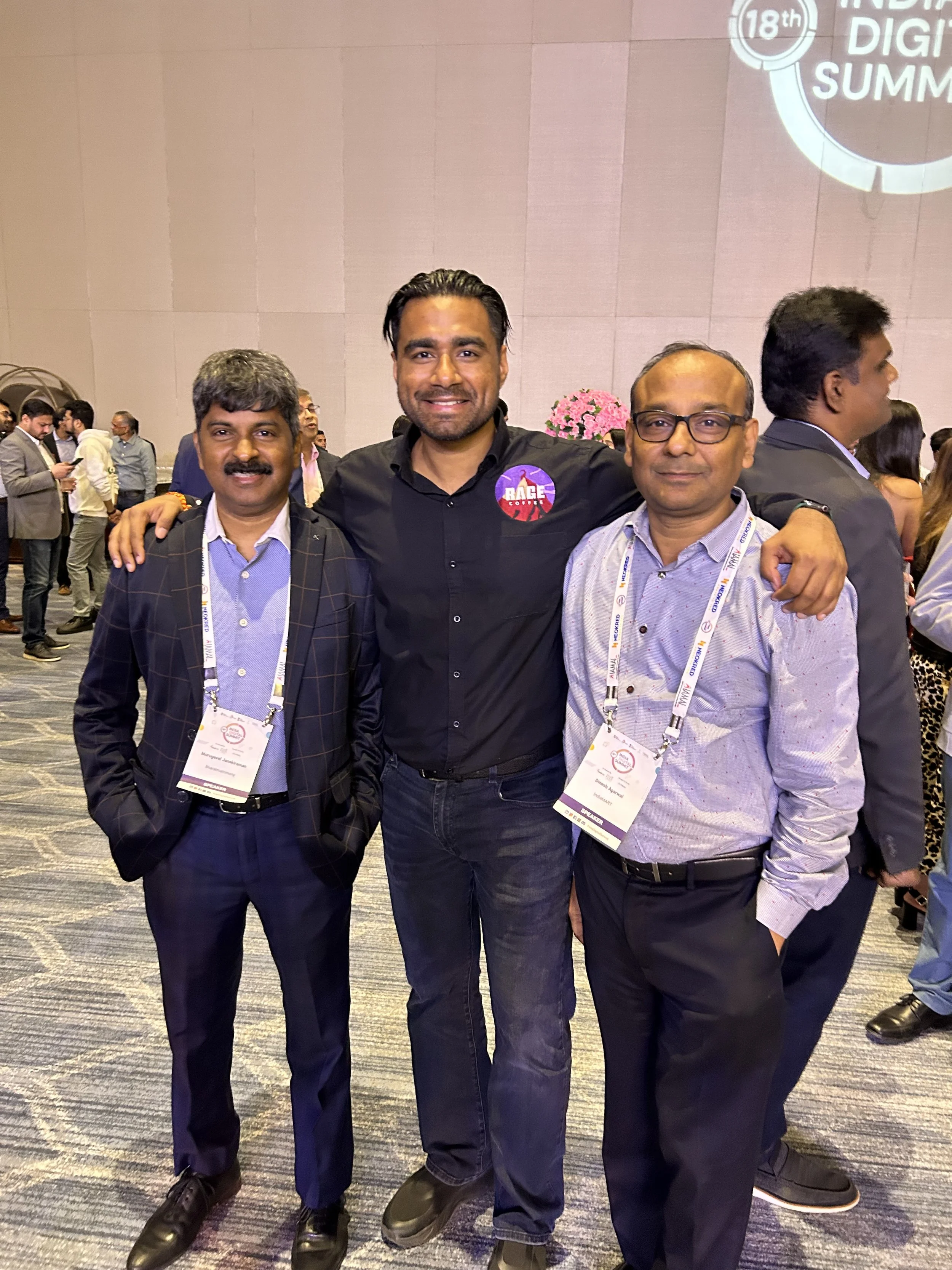 Three men standing inside a conference hall, smiling at the camera. The man in the middle has his arms around the other two men, all wearing business casual attire with conference badges.
