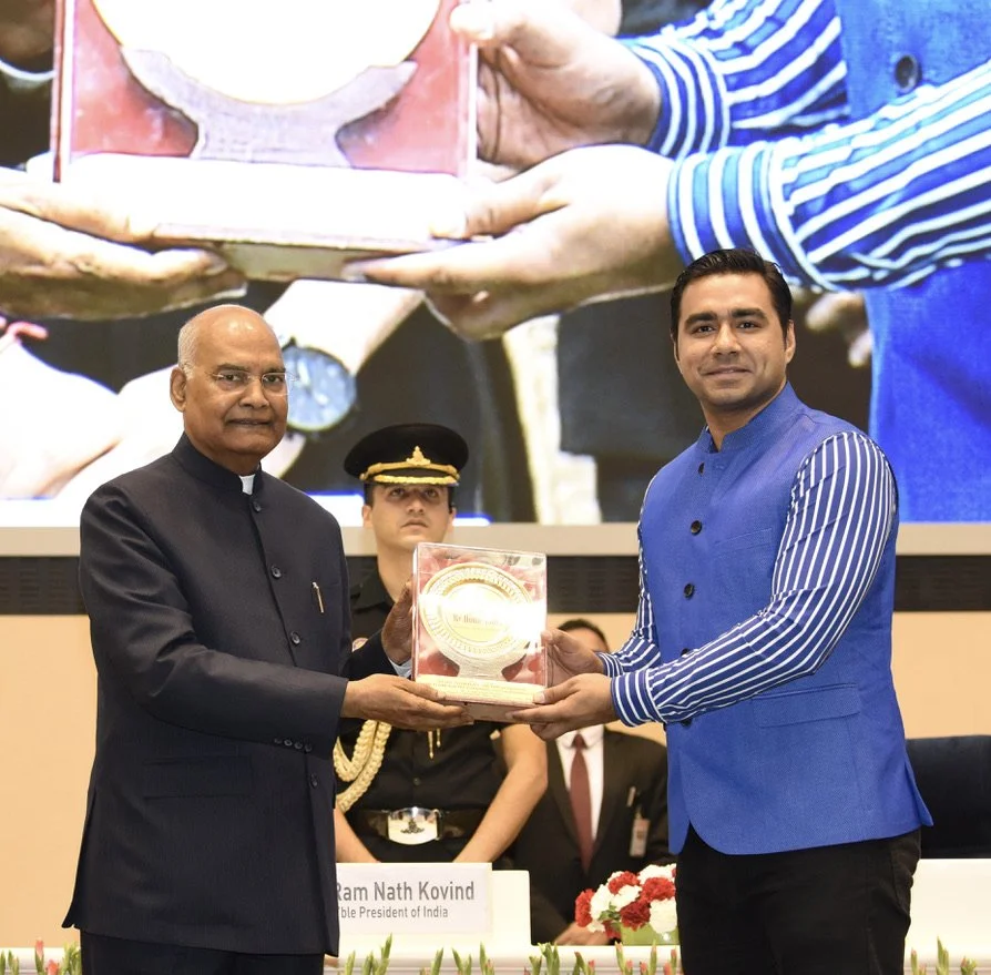Man in a black suit and glasses receiving an award or trophy from a man in a blue vest and striped shirt at a formal event. Several other individuals are standing in the background, including a woman in a uniform.