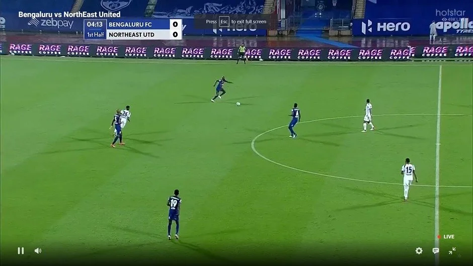 A soccer match between Bengaluru FC and NorthEast United. Players are on the field, with one player in blue chasing the ball. The scoreboard shows 0-0, with 4 minutes and 13 seconds elapsed in the first half. Advertising banners are visible around th