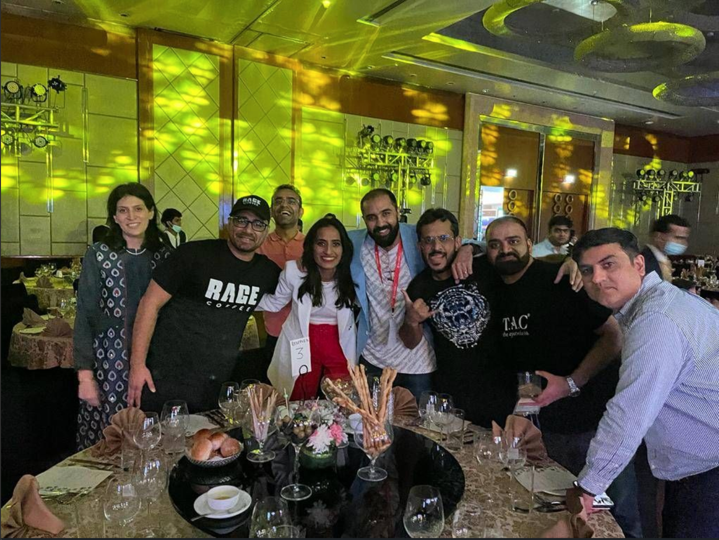 Group of eight people smiling and posing for a photo at a banquet table, celebrating in a decorated event hall with colorful lighting and flower arrangements.