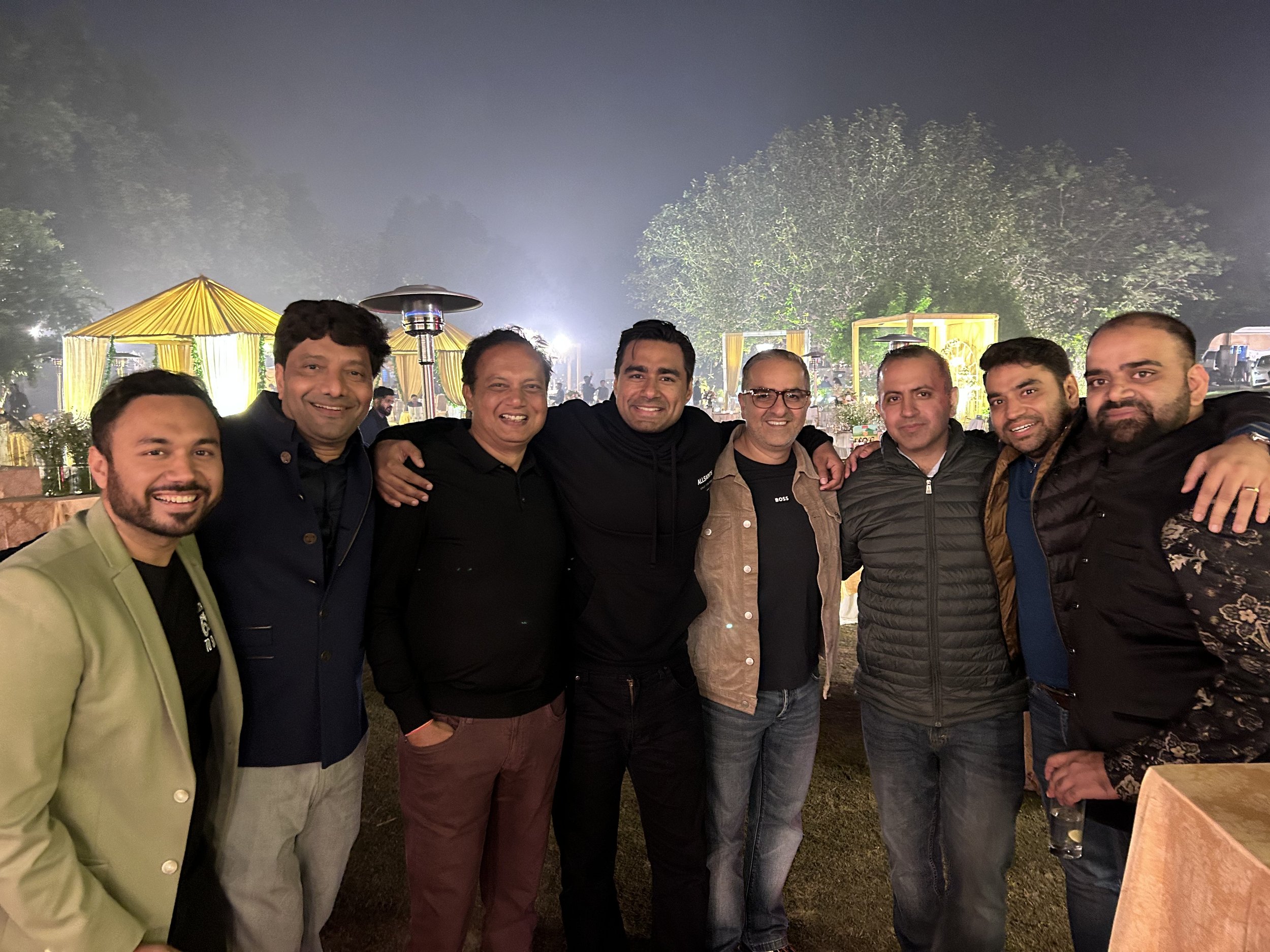 Group of nine men standing close together outdoors at night, smiling with arms around each other, in a festive setting with yellow tents and string lights in the background.