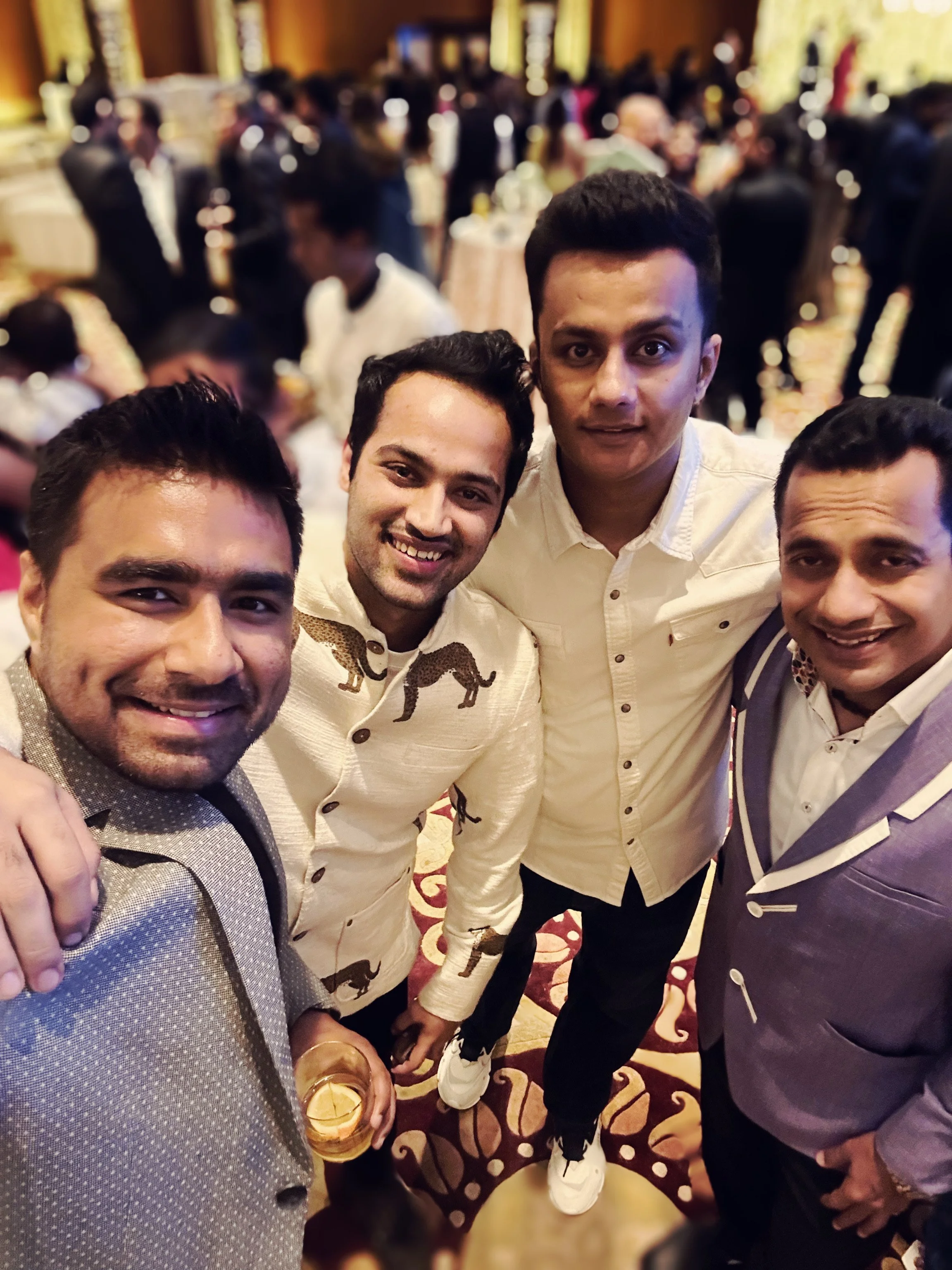 Four men posing for a photo at a lively event or party, with blurred guests and elegant decor in the background.