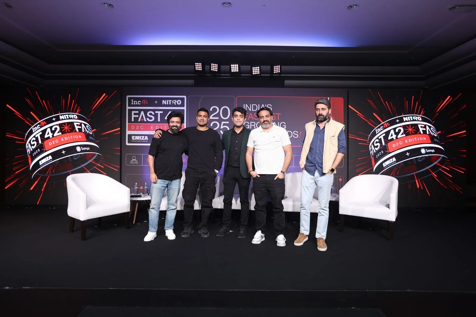 Five men standing on stage at a conference with a large digital display behind them. The display has a logo for the 'Fast 42' event, mentioning it is the 'D2C Edition 2025'. Two white chairs are positioned on either side of the men, with water bottle