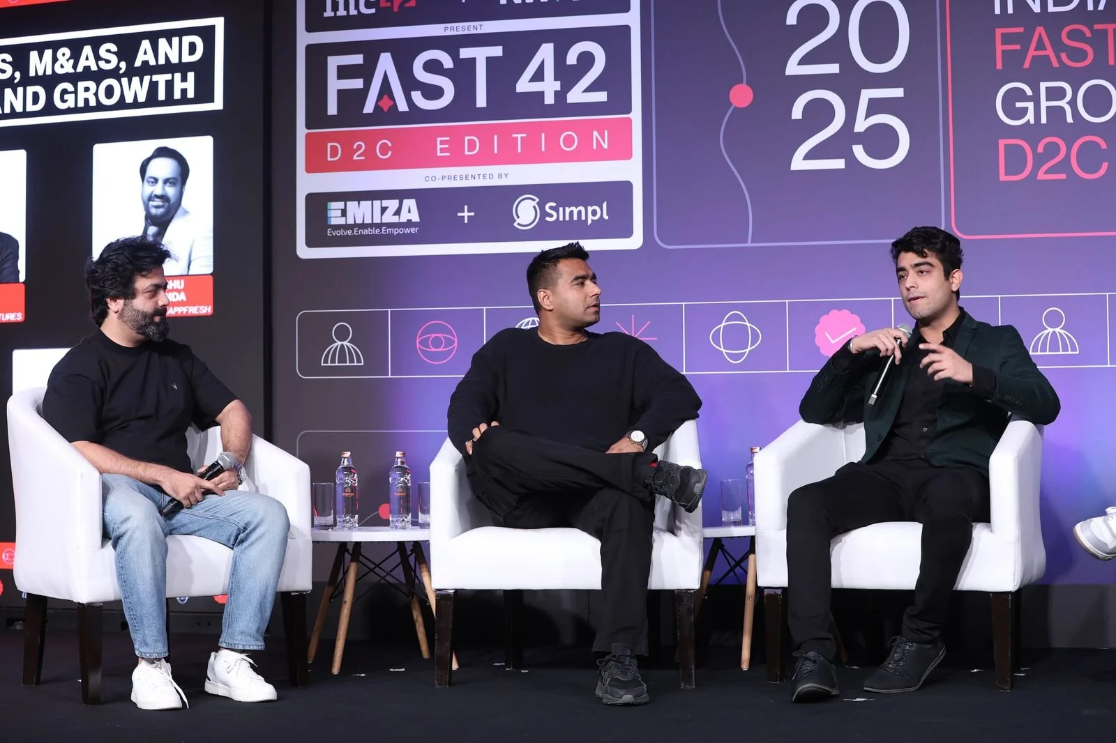 Three men sitting on stage during a panel discussion at an event titled 'Fast 42 D2C Edition' with a large digital display behind them. The display shows event details and logos, and three icons are visible below the display.