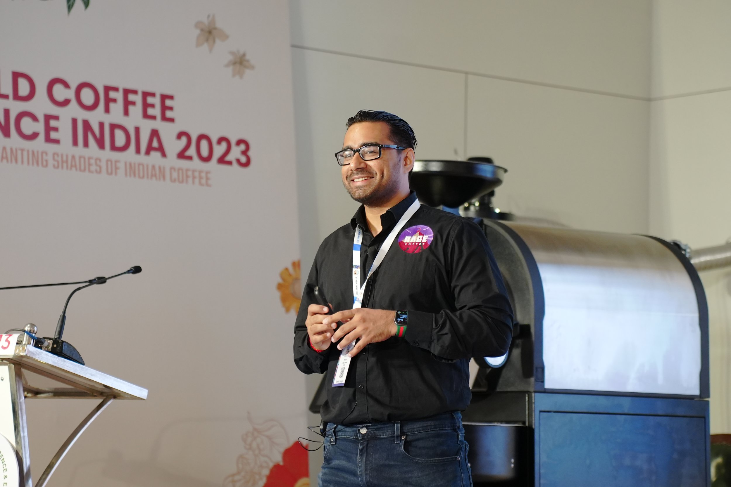 A man with glasses, smiling and wearing a black shirt with a “Race Coffee” badge, standing in front of a coffee roasting machine at the World Coffee Conference India 2023.