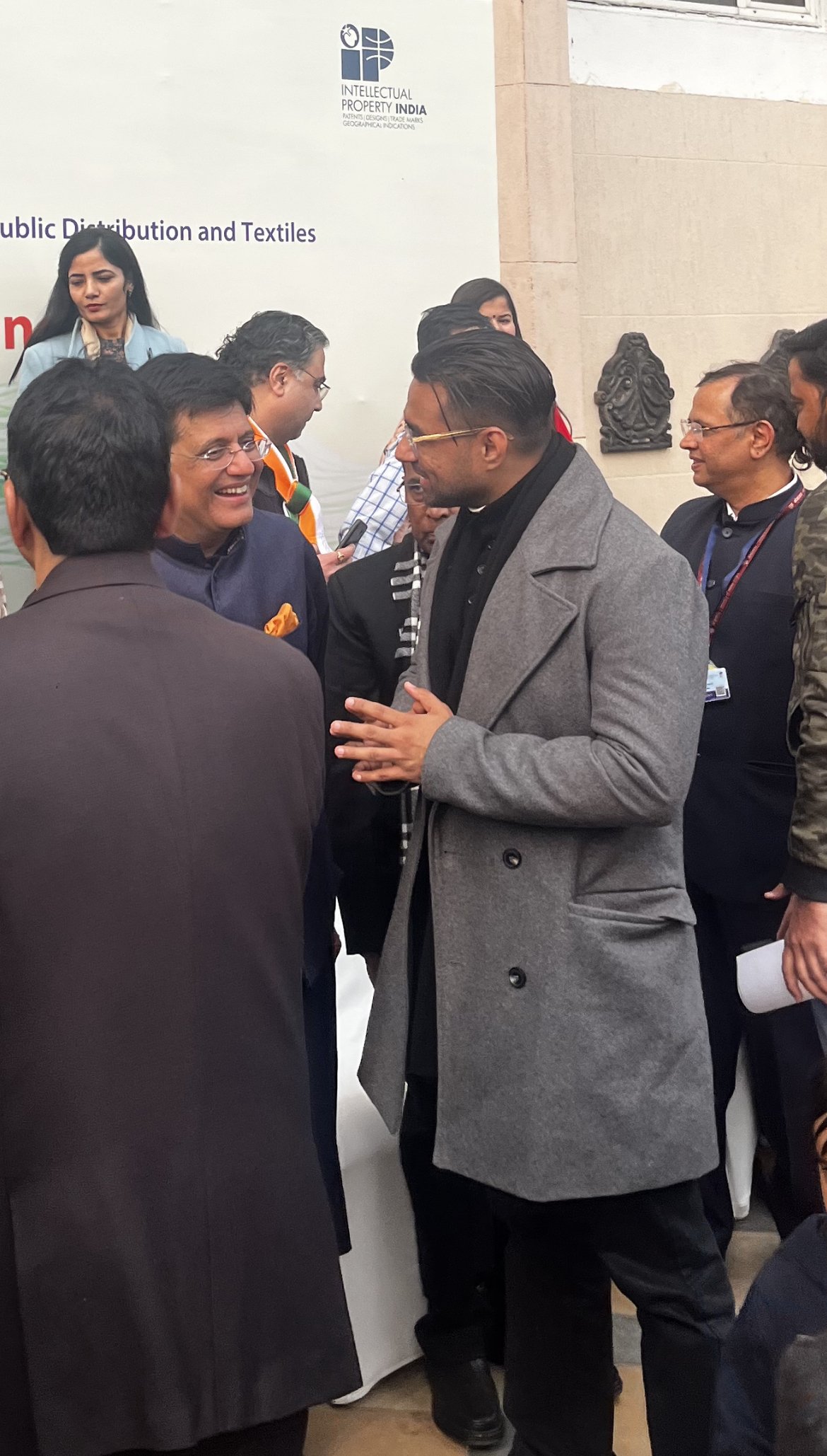 Group of people engaged in conversation at an event, with a man in a gray coat speaking and others smiling and listening, in front of a white wall with a sign about intellectual property India.