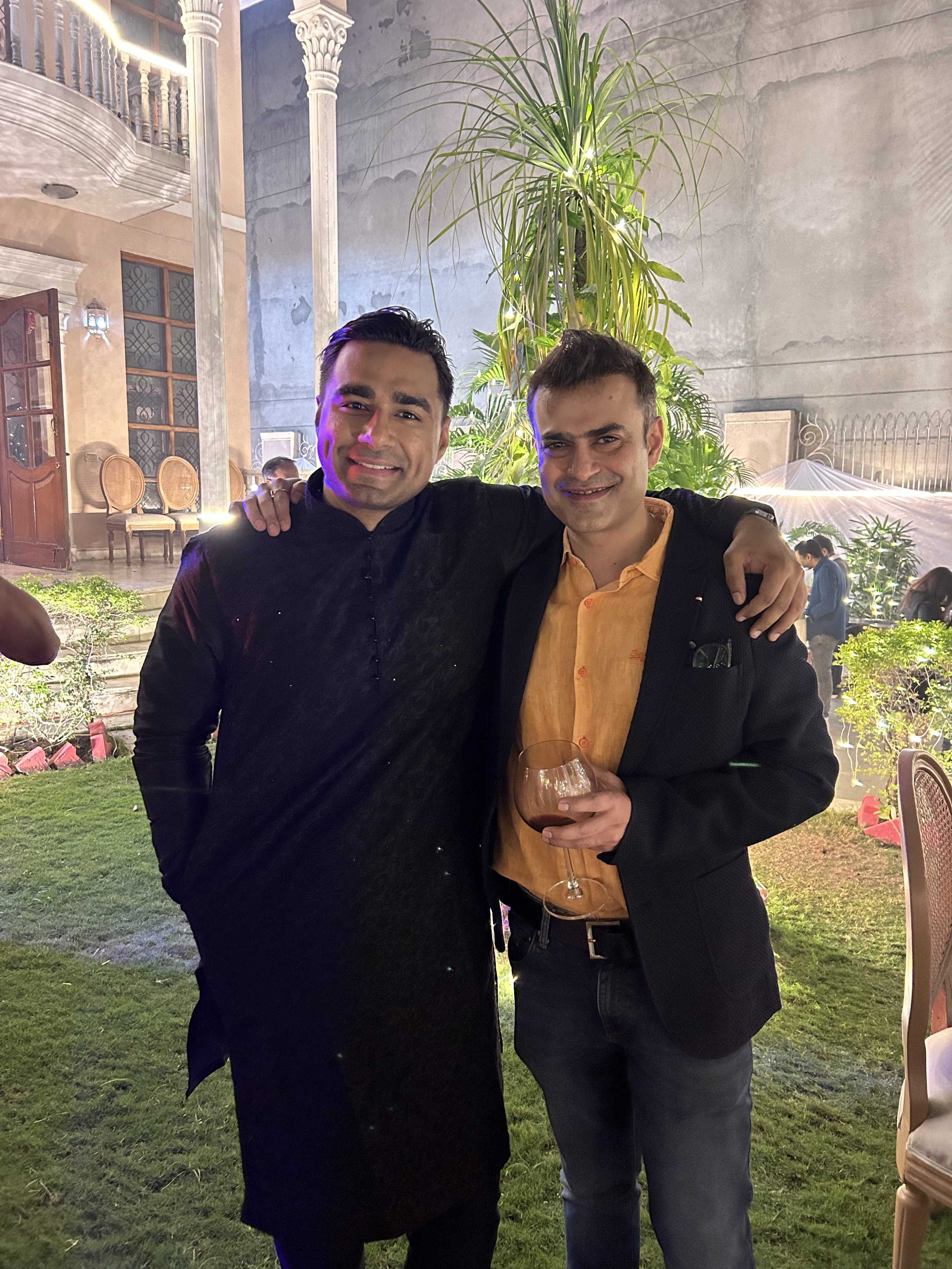 Two men standing together at an outdoor event during the evening, smiling at the camera. One man is wearing a black traditional outfit and the other is dressed in a black blazer with a yellow shirt, holding a glass of wine. In the background, there i