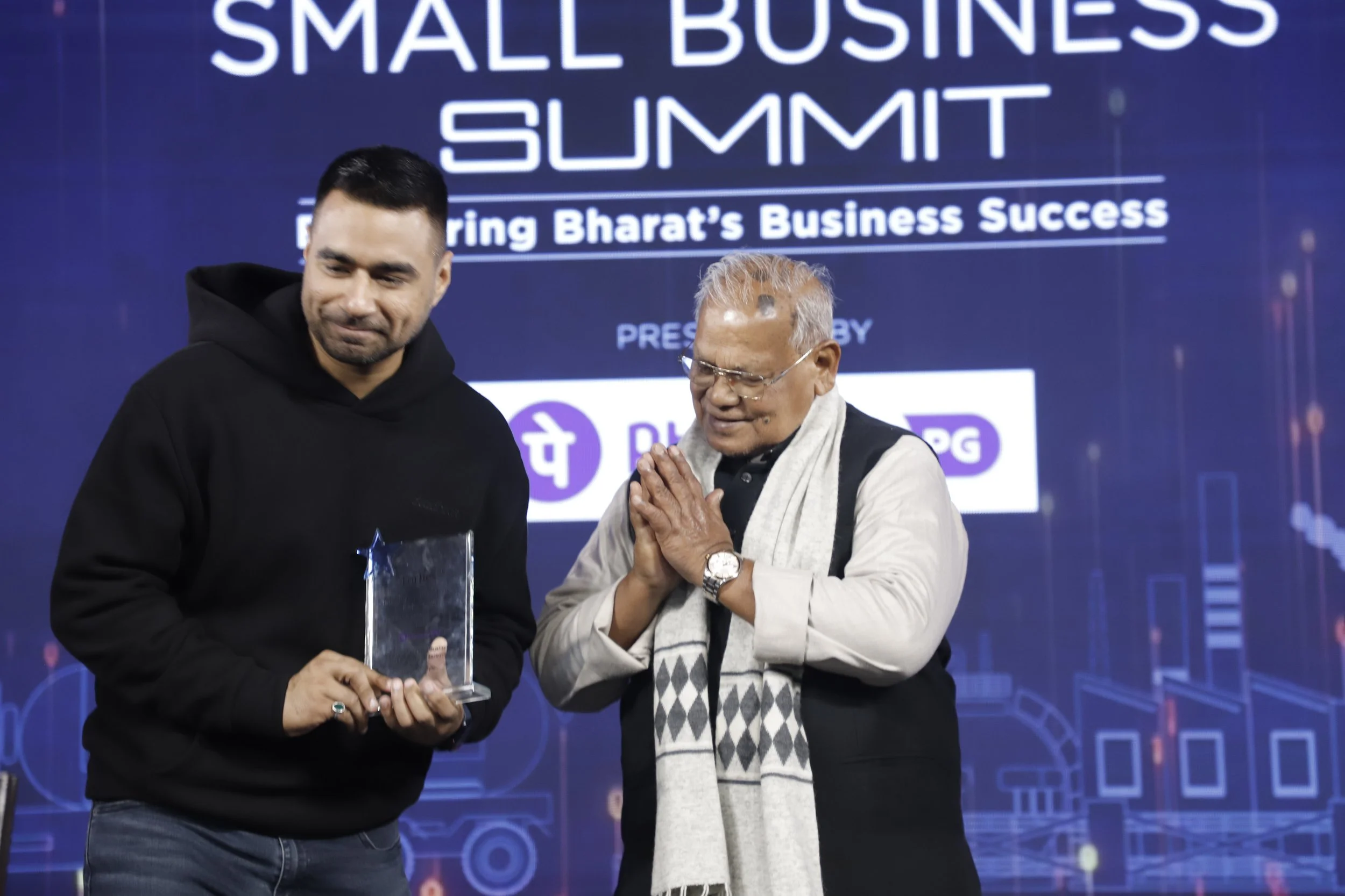 Two men on stage at the Small Business Summit, one holding an award trophy, with a large screen in the background displaying event details and logos.