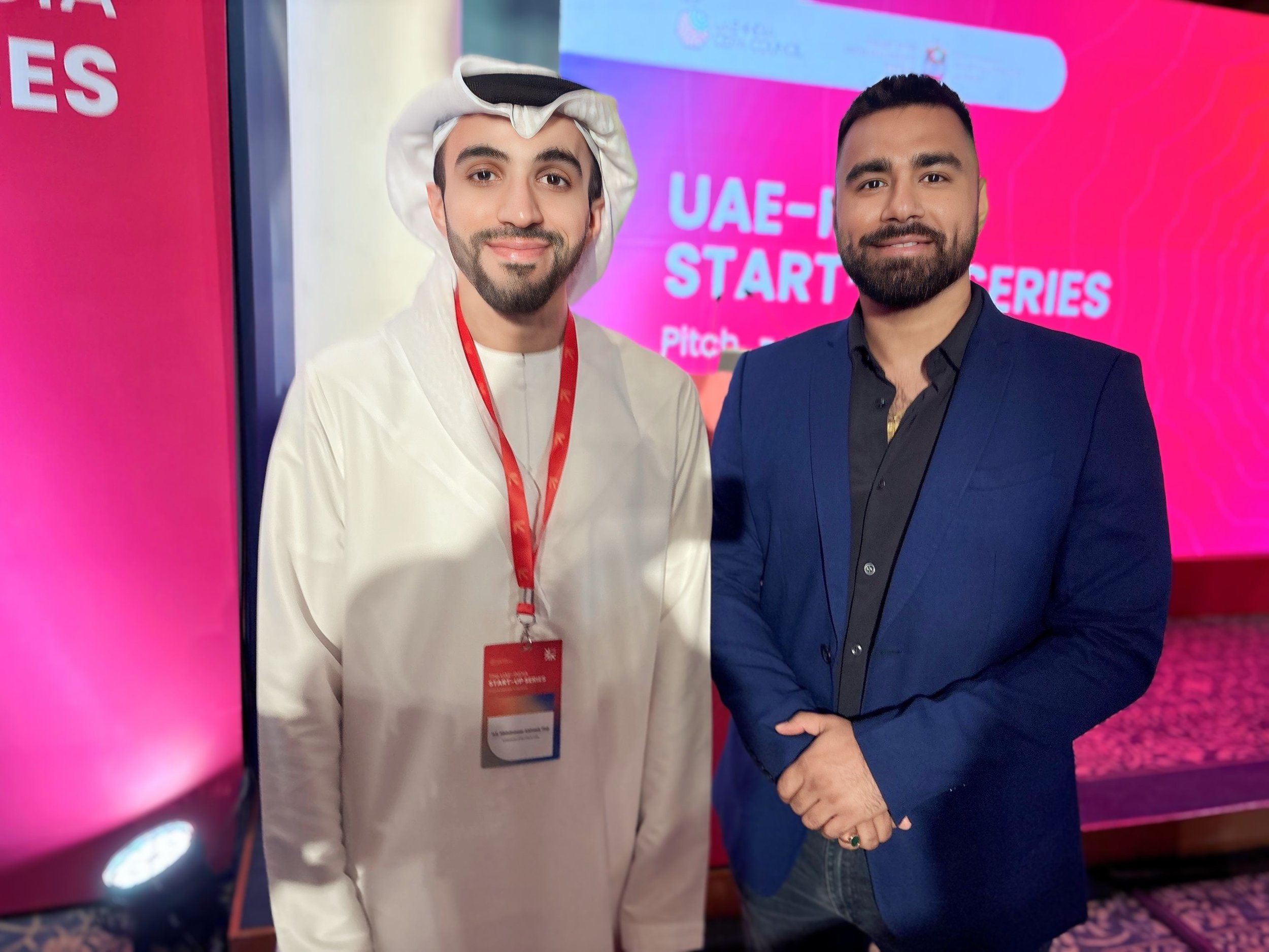 Two men standing together at a startup event with a pink and purple background displaying 'UAE-I STARTUP SERIES' and 'Pitch'. One man is wearing traditional Middle Eastern attire with a white thobe and head covering, red lanyard, and name badge. The 