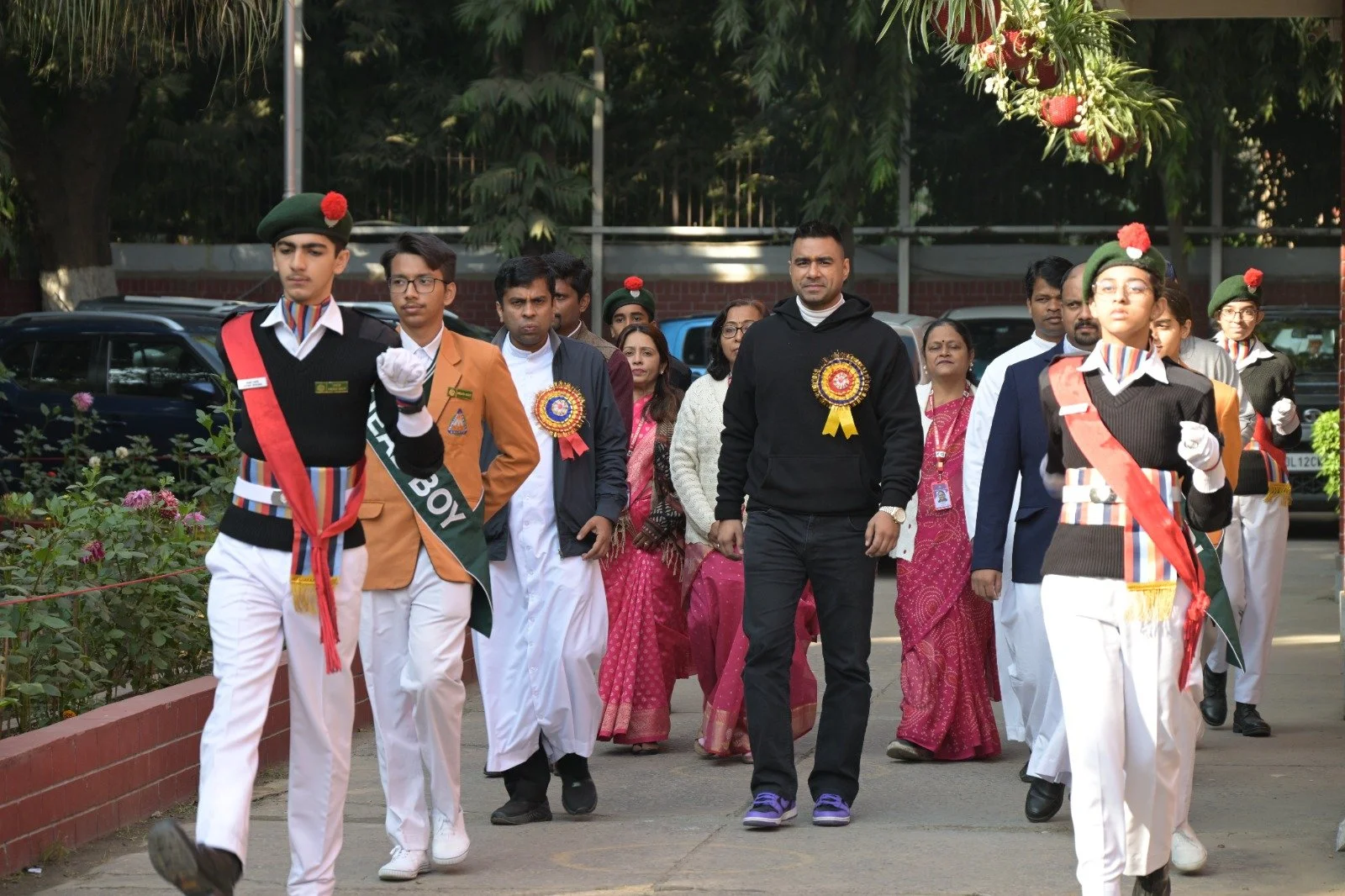 Group of people participating in a parade or ceremony, including students in uniform and officials, walking on a sidewalk with trees and parked cars in the background.