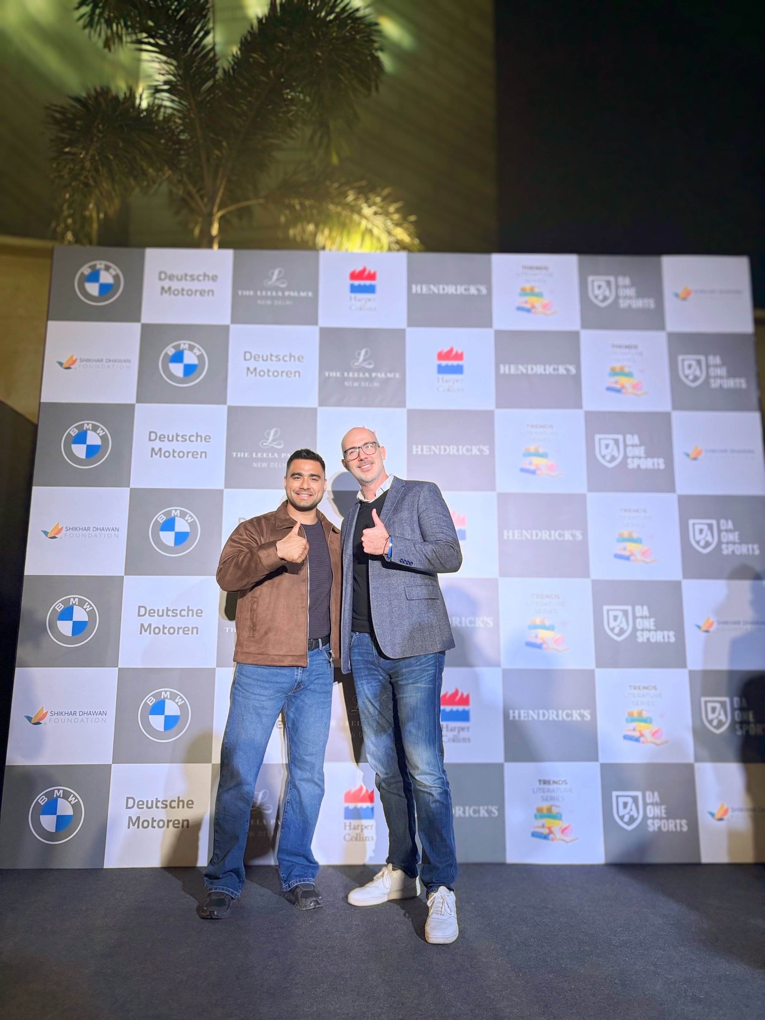Two men standing together at an event, smiling and giving thumbs up in front of a step and repeat banner with logos of Deutsche Motoren, BMW, Hendrick's, Harper Collins, and other sponsors, with a palm tree and outdoor night sky in the background.