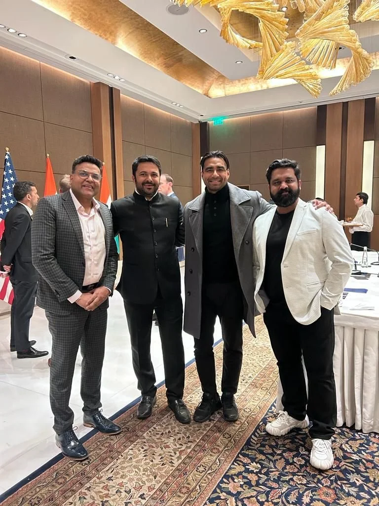 Four men standing together, dressed in formal and semi-formal attire, smiling for a photo in a conference room with flags and other people in the background.