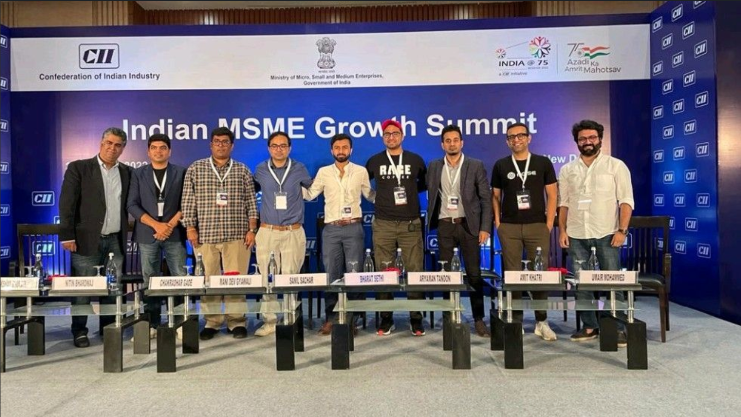 A group of nine men standing on stage at the Indian MSME Growth Summit, with a blue backdrop displaying logos and event details.