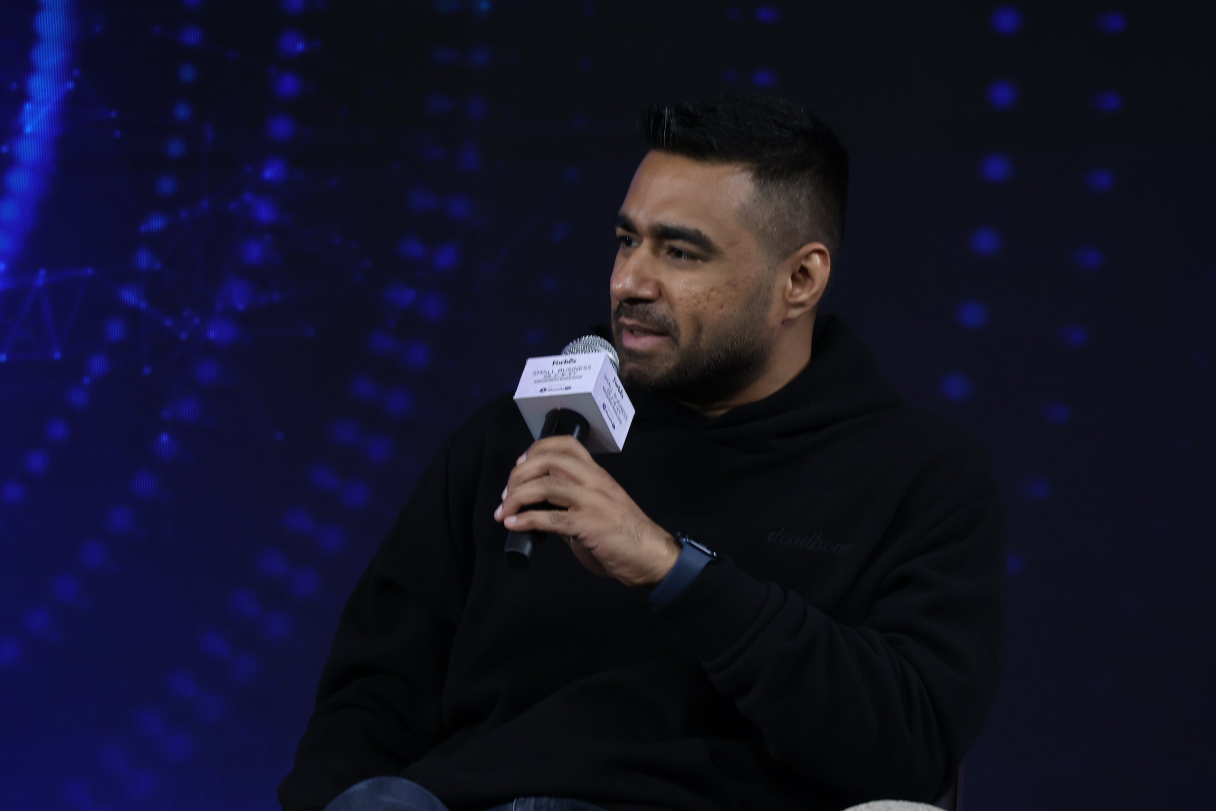 A man holding a microphone, speaking during an event, with a dark background featuring blue digital patterns.