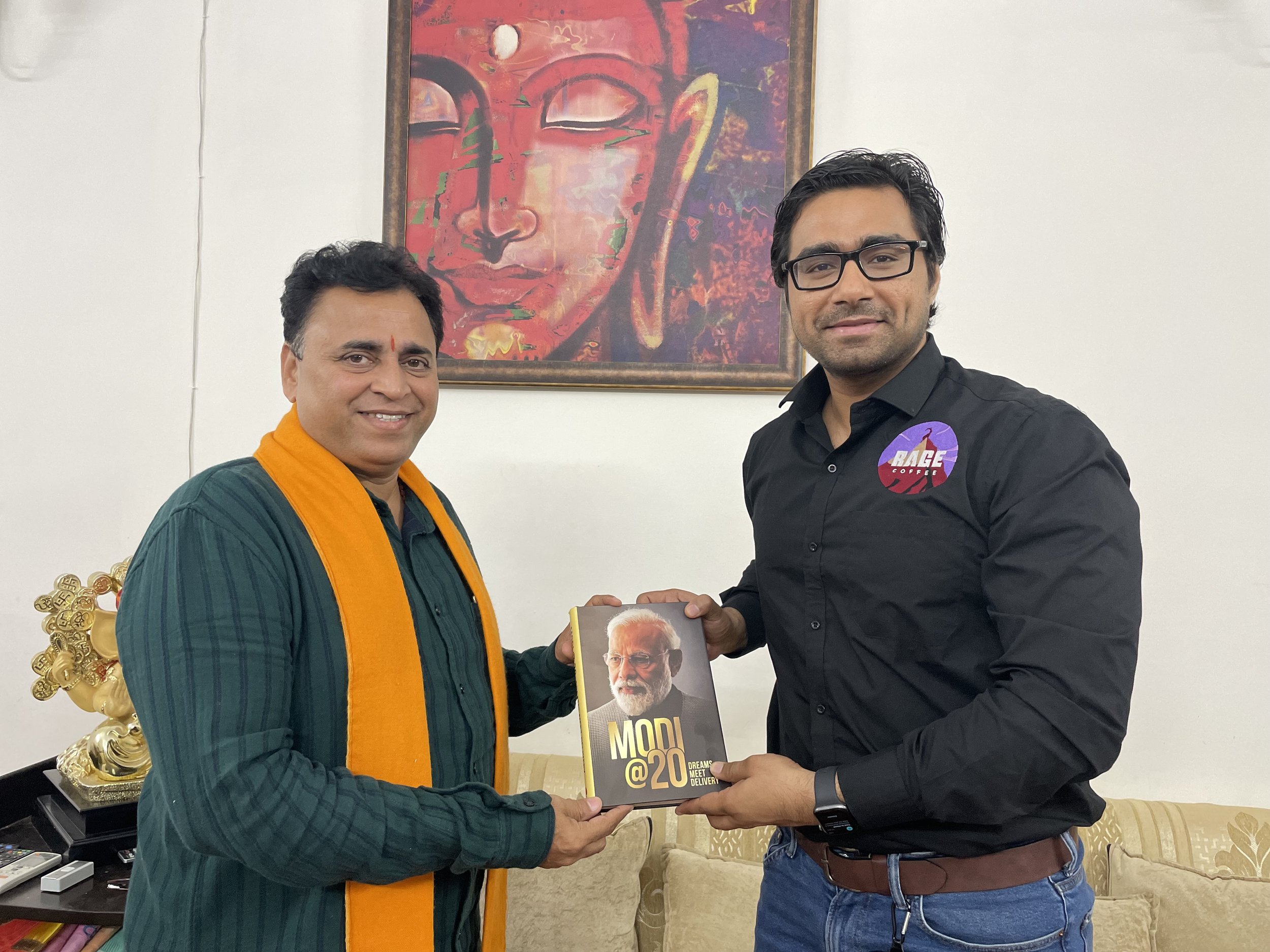 Two men standing in a living room, exchanging a book. The man on the left wears a green shirt with a yellow scarf, and the man on the right wears glasses and a black shirt with a badge that reads 'RACE.' There is a large framed painting of a Buddha f