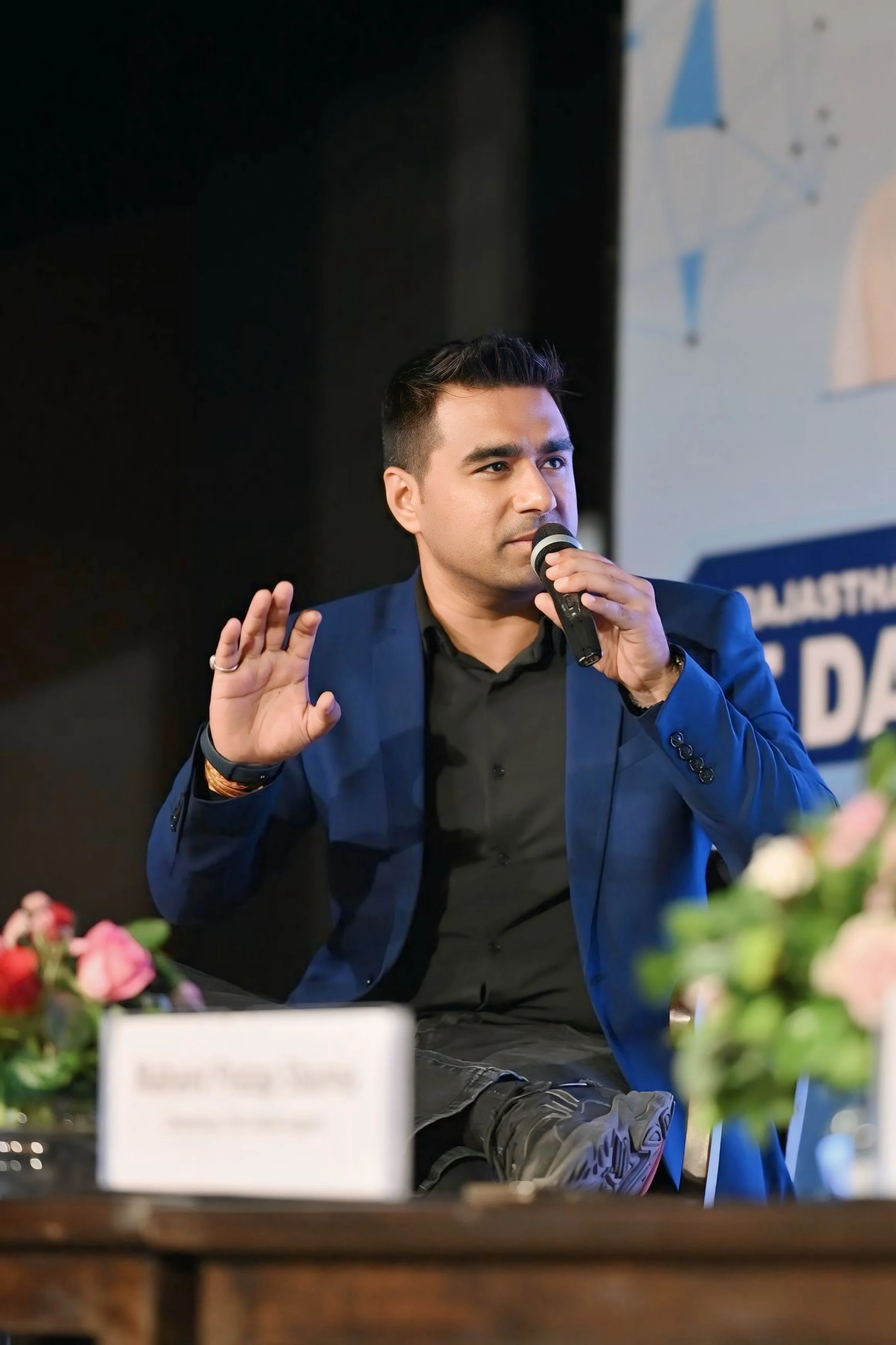 A man in a blue blazer and black shirt sitting on a stage, holding a microphone, raising his left hand with an open palm, speaking during an event or panel discussion.