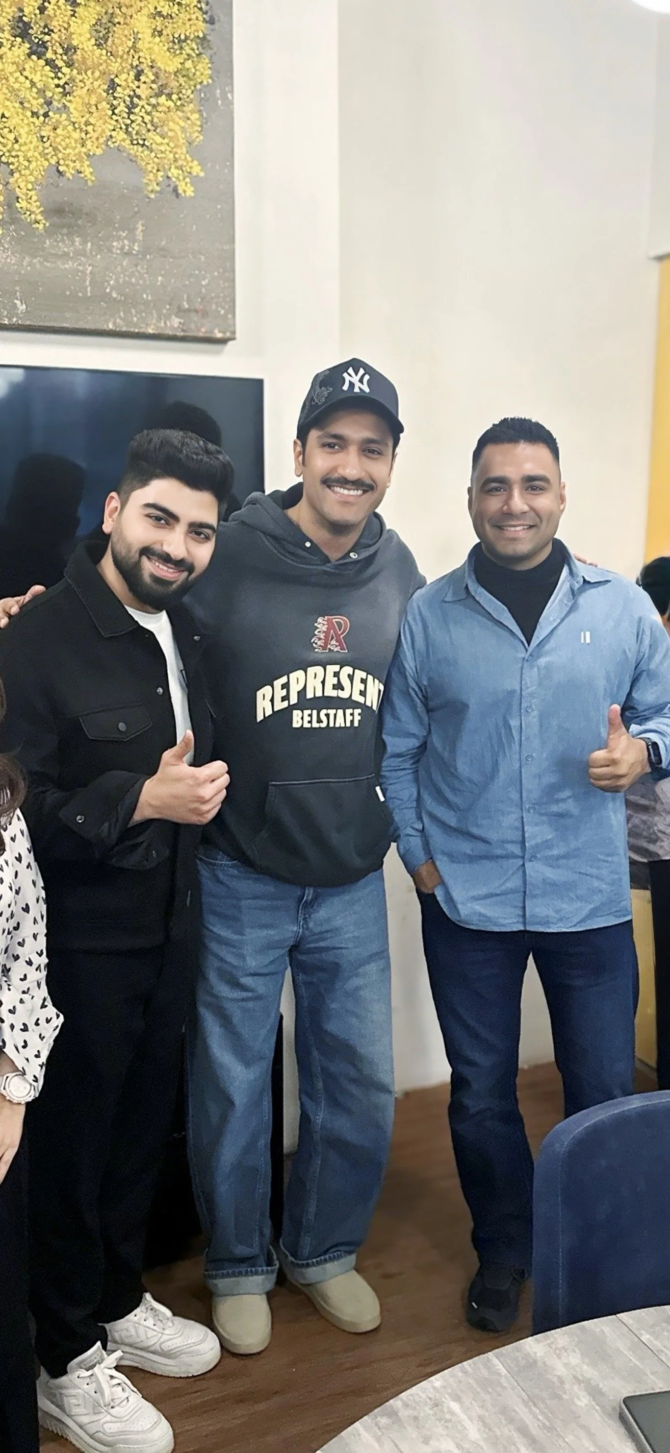 Three men standing together inside a room, smiling and showing thumbs up, with a television and a yellow wall in the background.