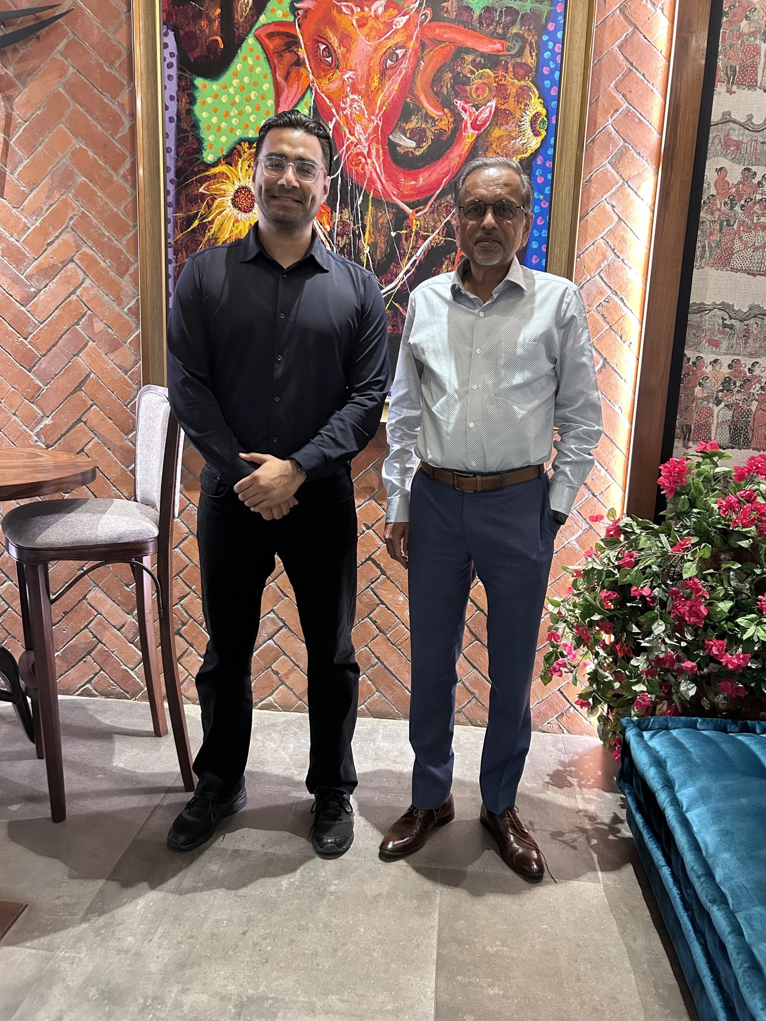 Two men standing indoors in front of a colorful painting, with brick and wooden wall decor, a small table with a chair, and a flowering plant nearby.