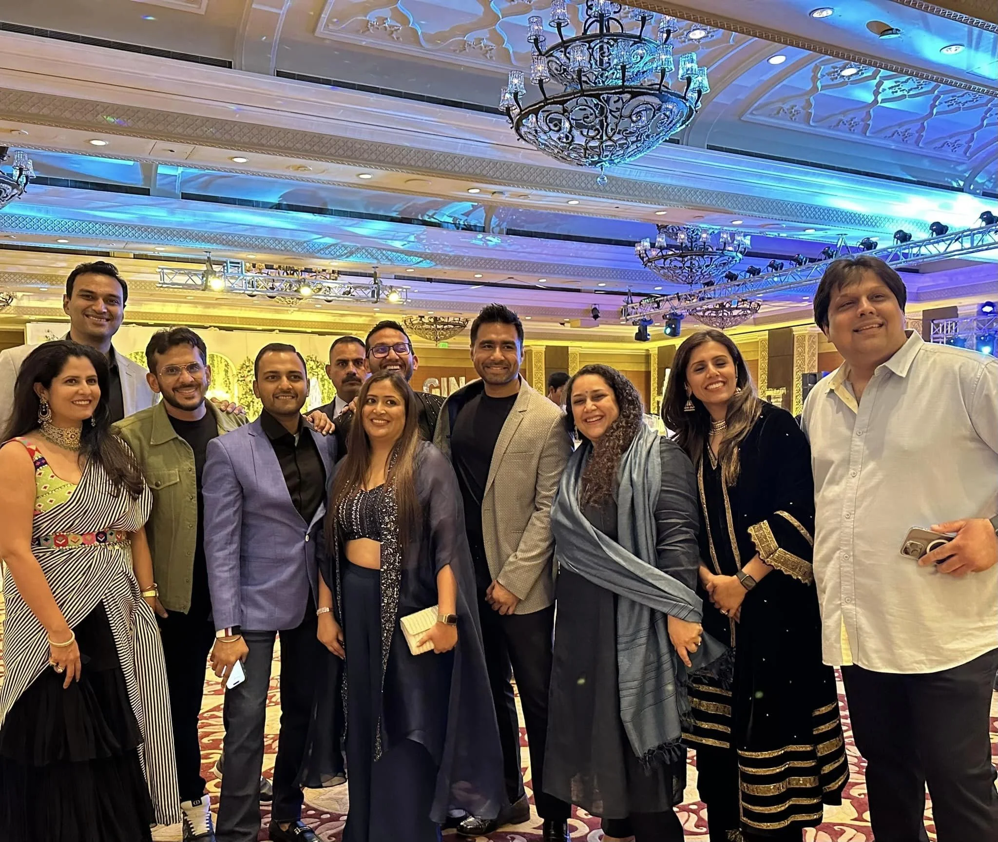 Group of ten people dressed in formal attire posing together at an indoor event with elaborate chandeliers and colorful lighting.