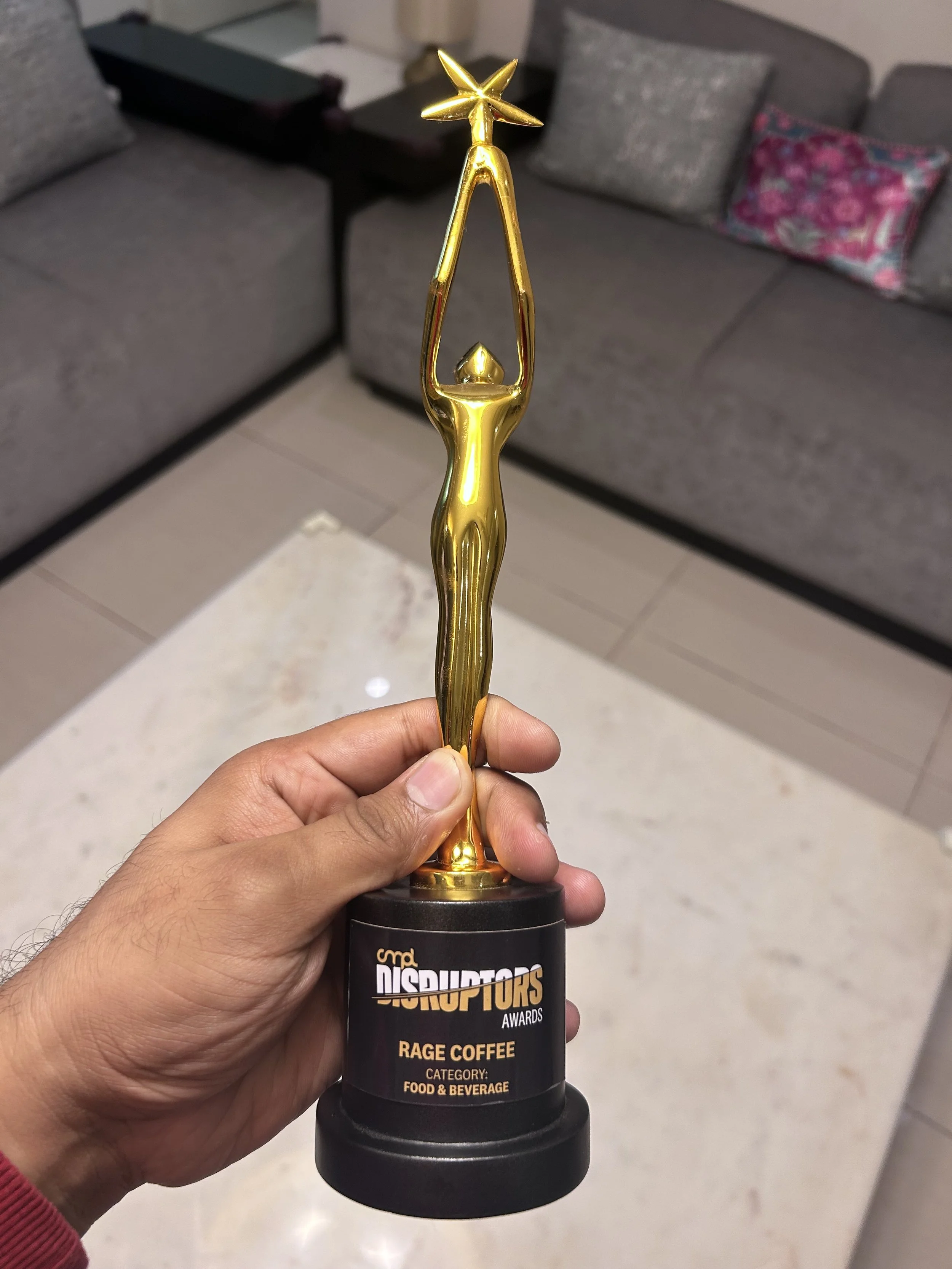 A person's hand holding a golden trophy with a humanoid figure reaching upward towards a star, placed on a black base with a label that reads 'Disruptors Awards - Rage Coffee - Category: Food & Beverage'. In the background, there are gray sofas and a