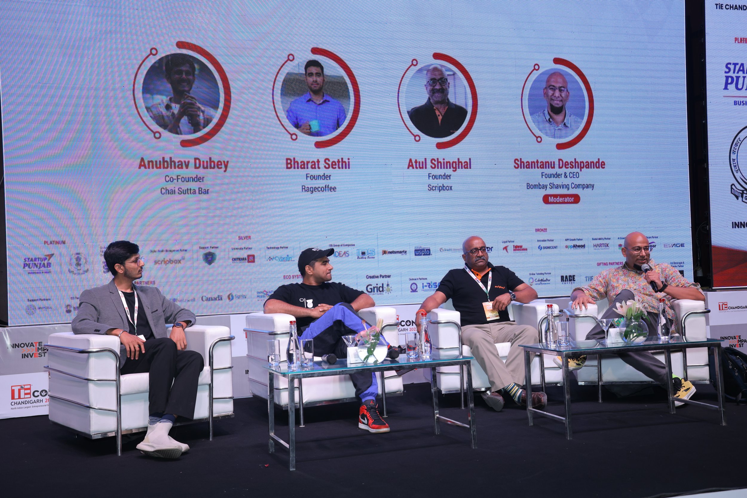 Panel discussion at a conference with five seated men on stage. Behind them, a large screen displays names and titles of four speakers, including Anubhav Dubey, Bharat Sethi, Atul Shinghal, and Shantanu Deshpande, with their respective roles and comp