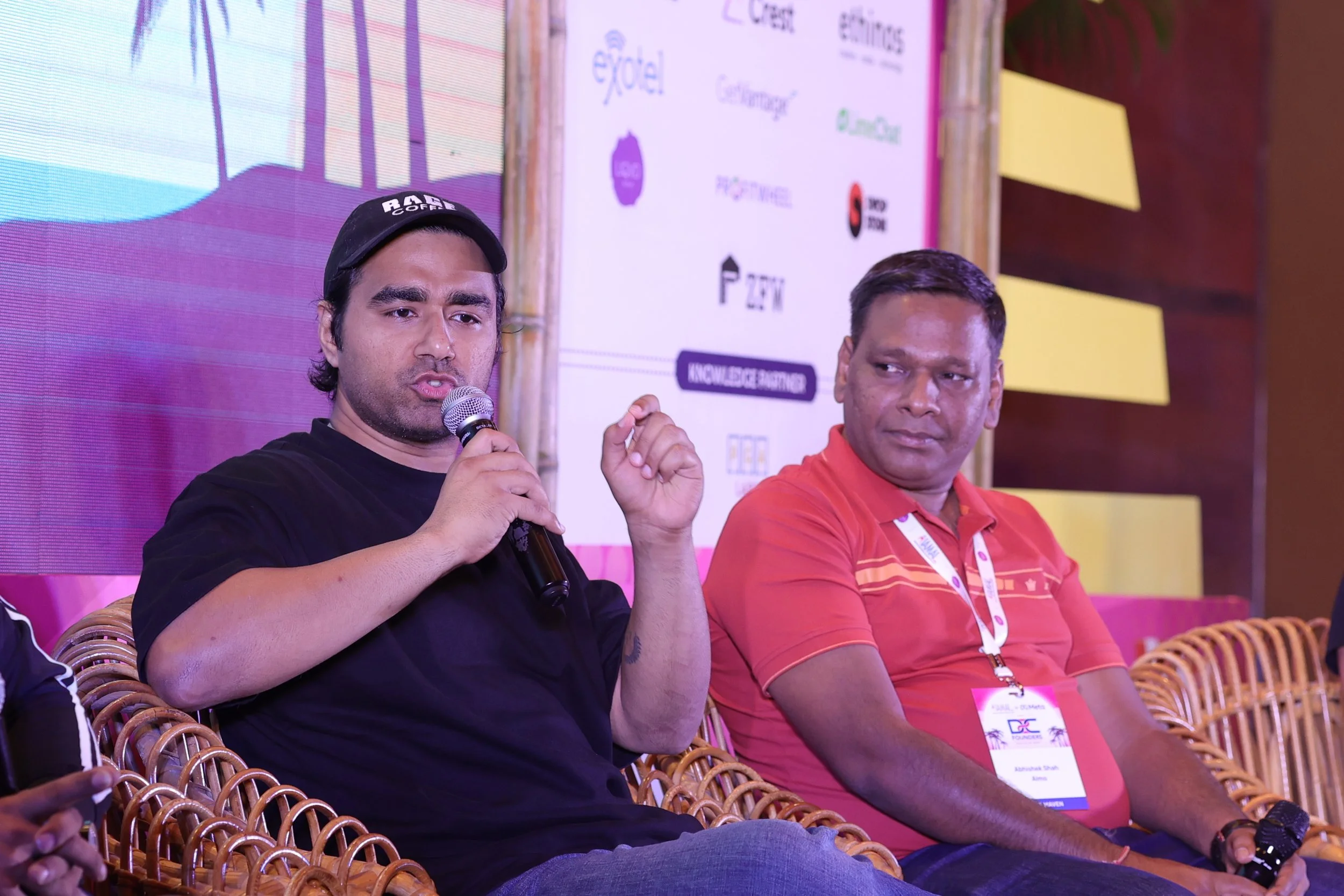 Two men sitting on stage during a conference, one speaking into a microphone and the other listening. The man on the left is wearing a black T-shirt and a cap, while the man on the right is in a red polo shirt with a conference badge.